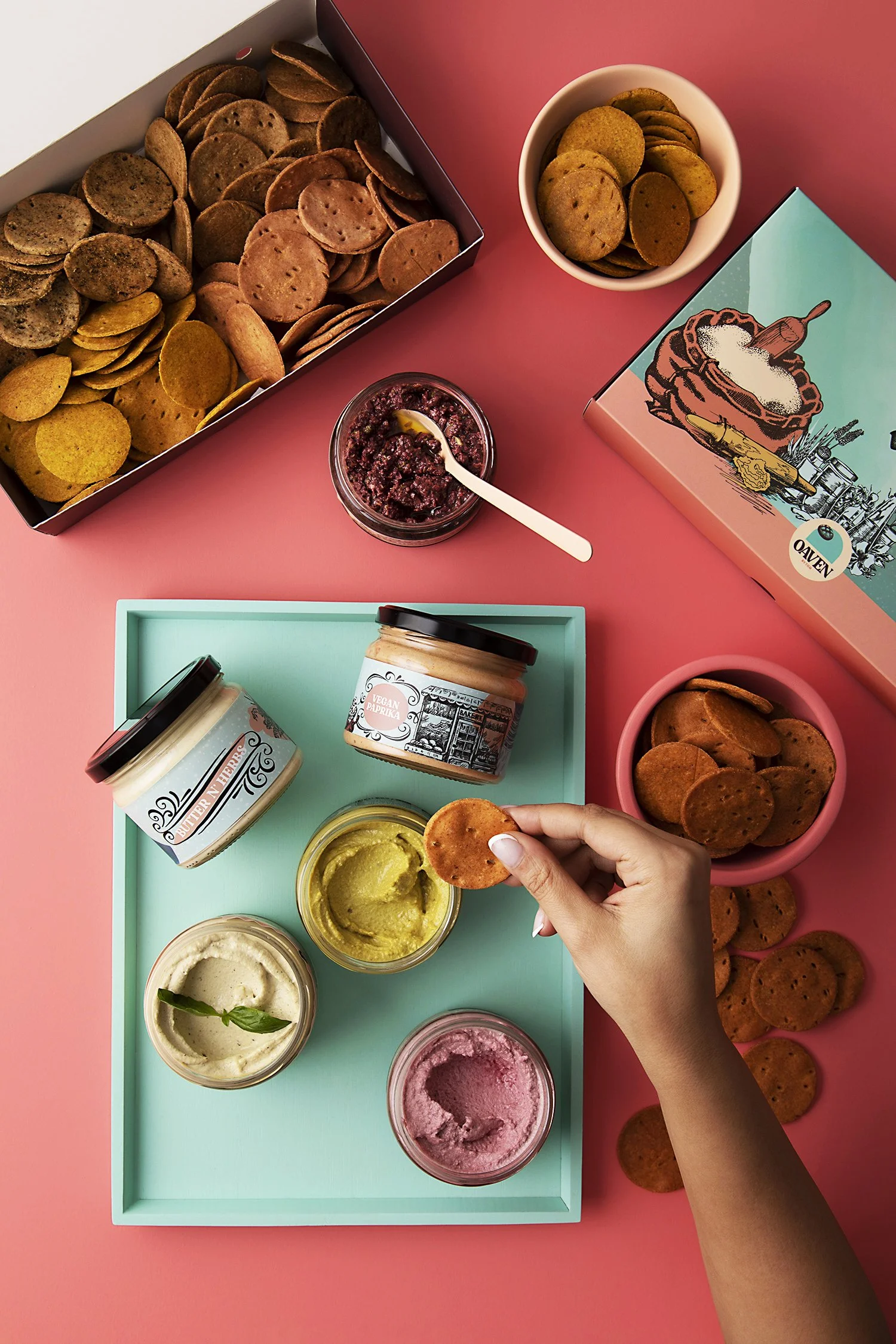 A colorful flat lay of snack items including bowls of crackers, various jars of dips or spreads, and ice cream or sorbet in small jars on a pink surface. 