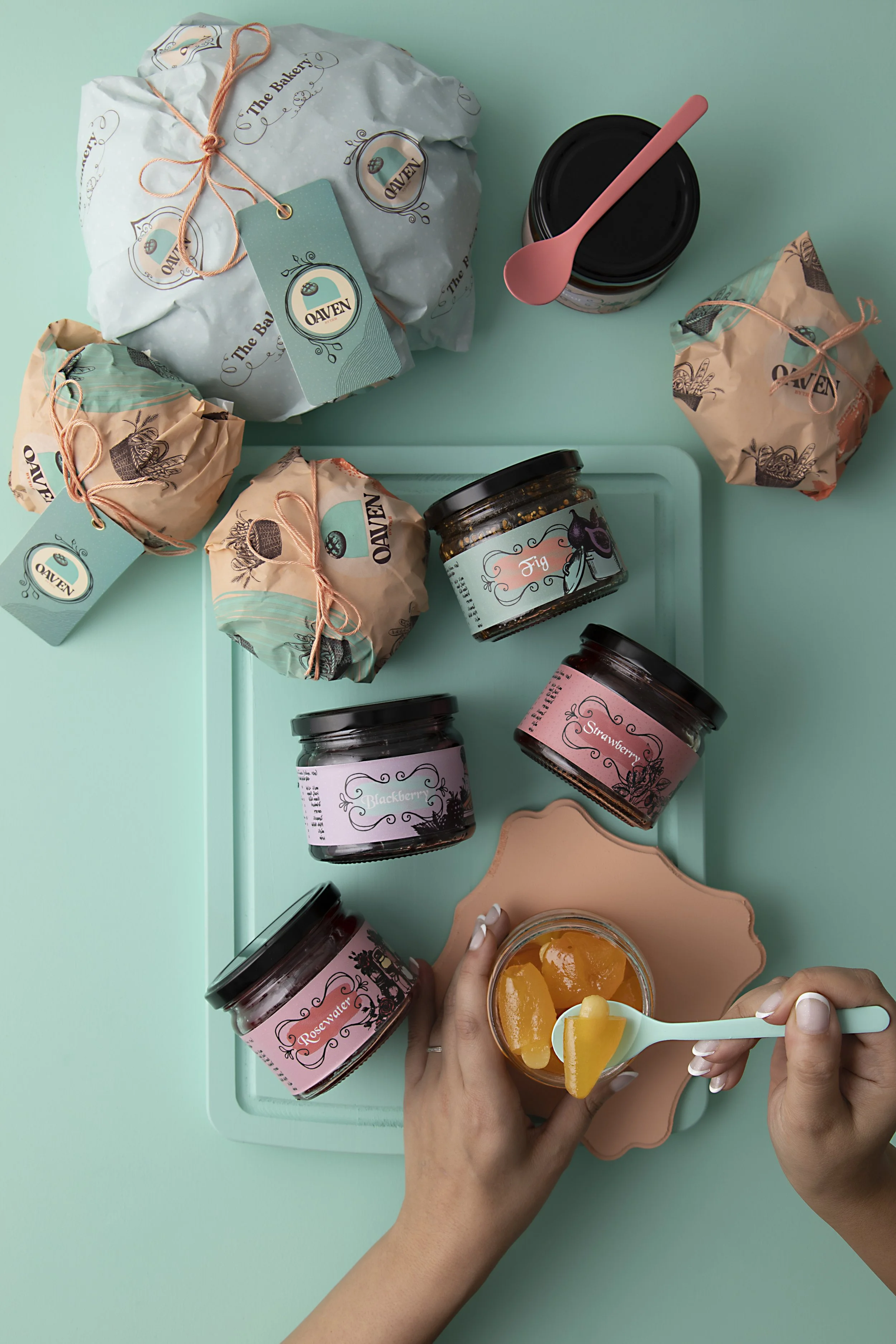 Assorted jars of jam and wrapped baked goods on a pastel turquoise surface, with a hand holding a jar of yellow jam and a spoon.