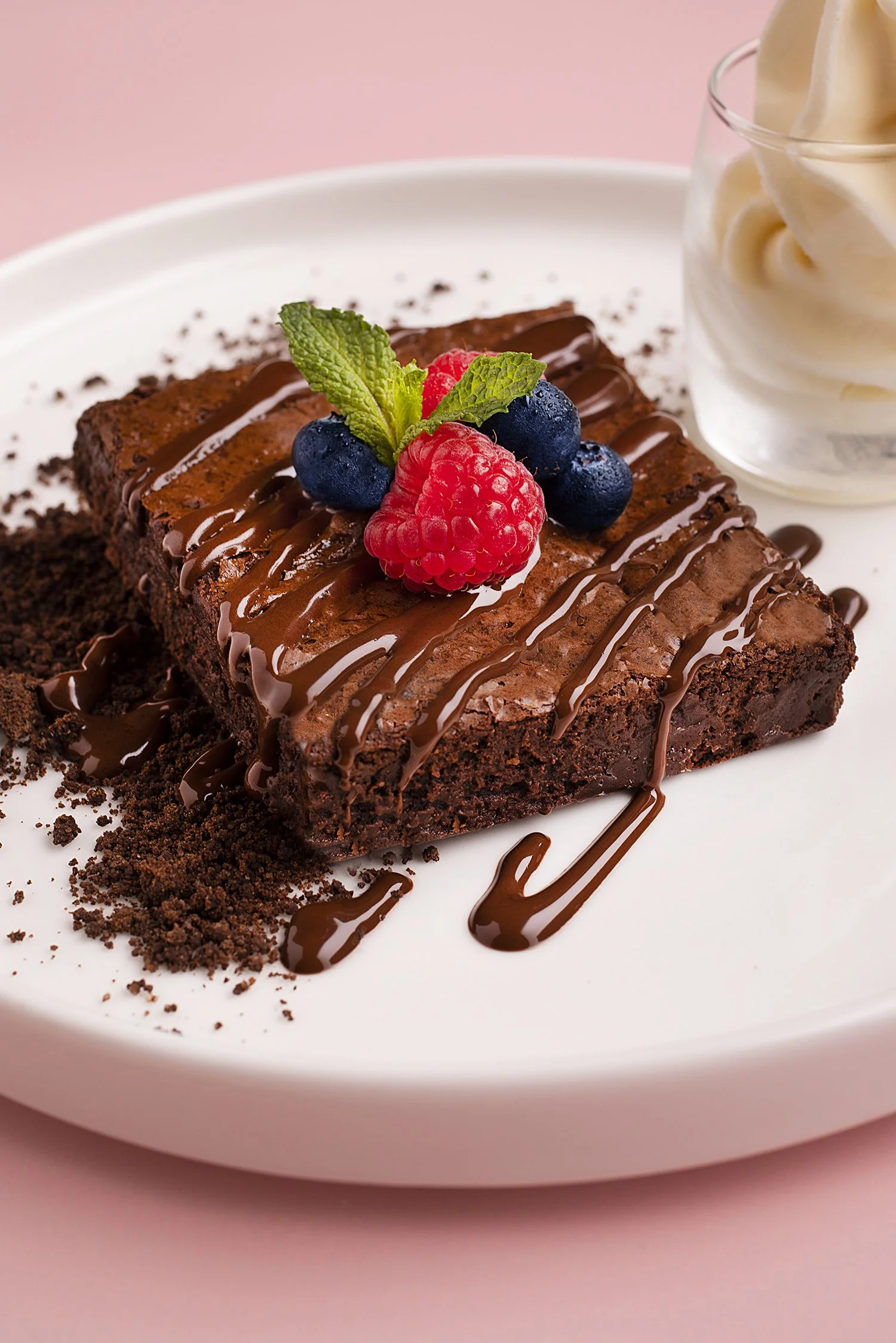 A slice of chocolate brownie topped with chocolate drizzle, fresh raspberries, blueberries, and mint leaves