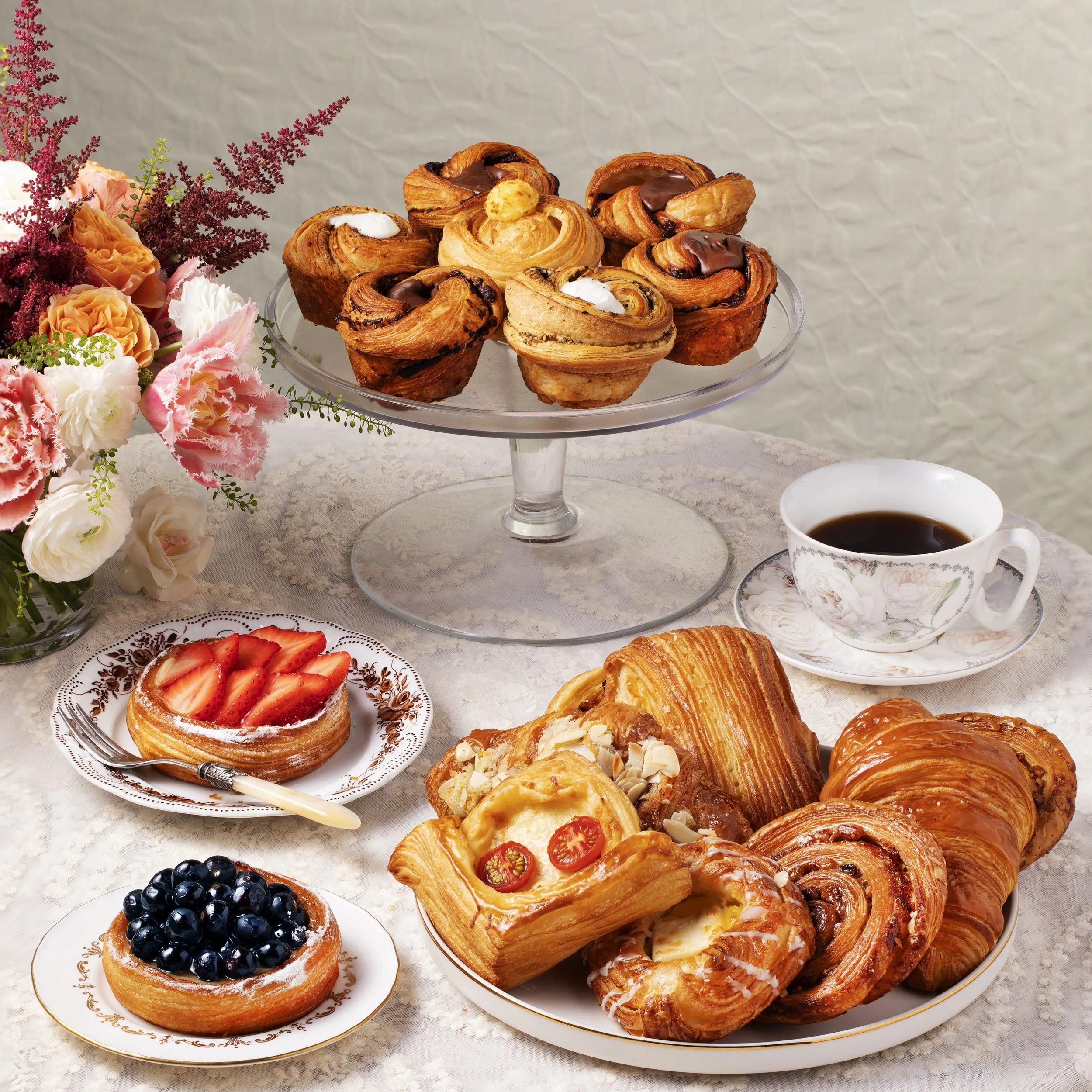A table with an assortment of pastries, including croissants, fruit tarts, and Danish pastries, a cup of coffee