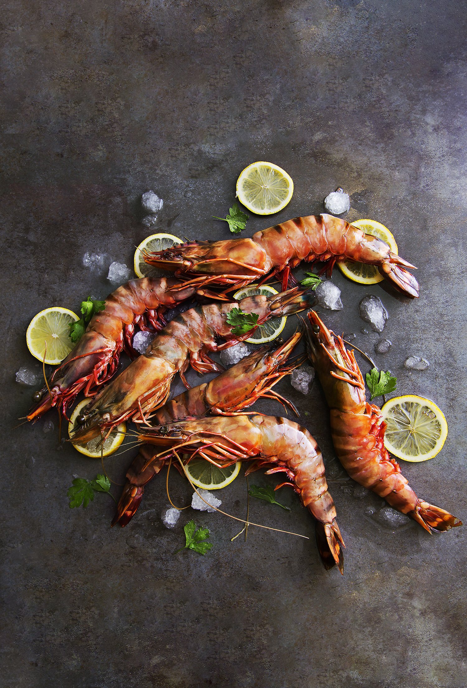 Fresh cooked shrimp garnished with lemon slices, parsley, and ice cubes on a dark surface. 