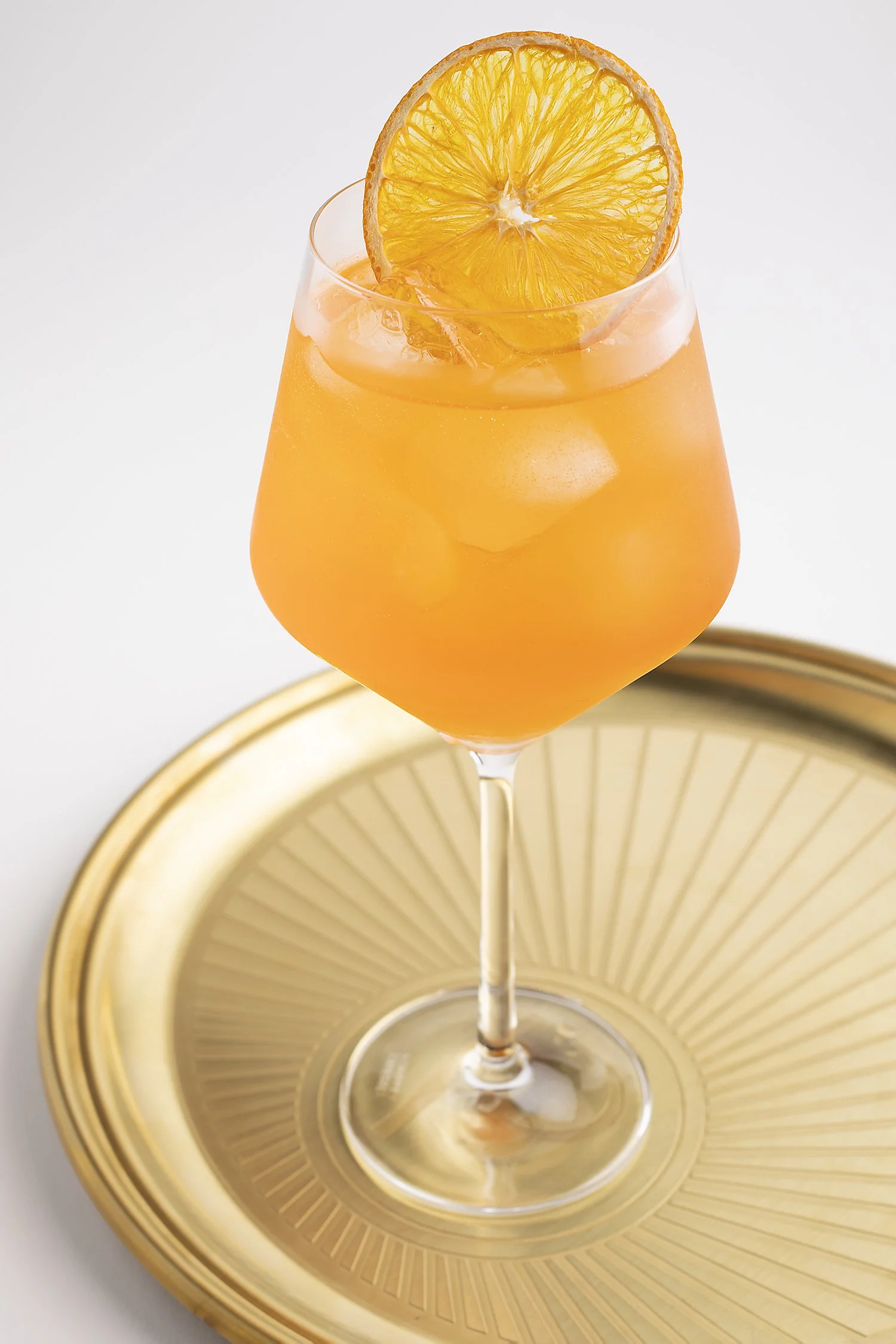 An orange-colored cocktail with ice cubes, garnished with a slice of orange, on a gold plate. 