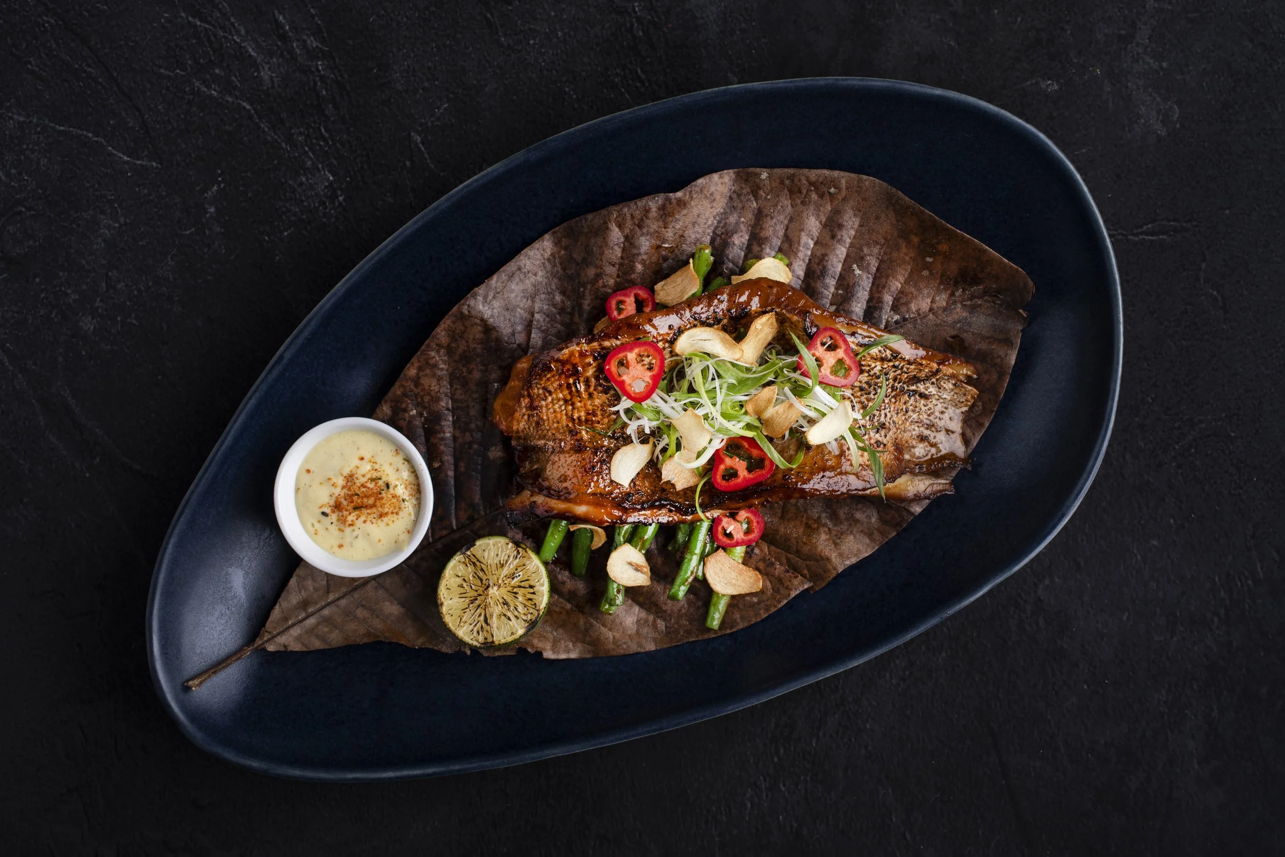 Grilled fish served on a black oval plate with grilled lemon, garnished with chili slices, green beans, and fried garlic chips