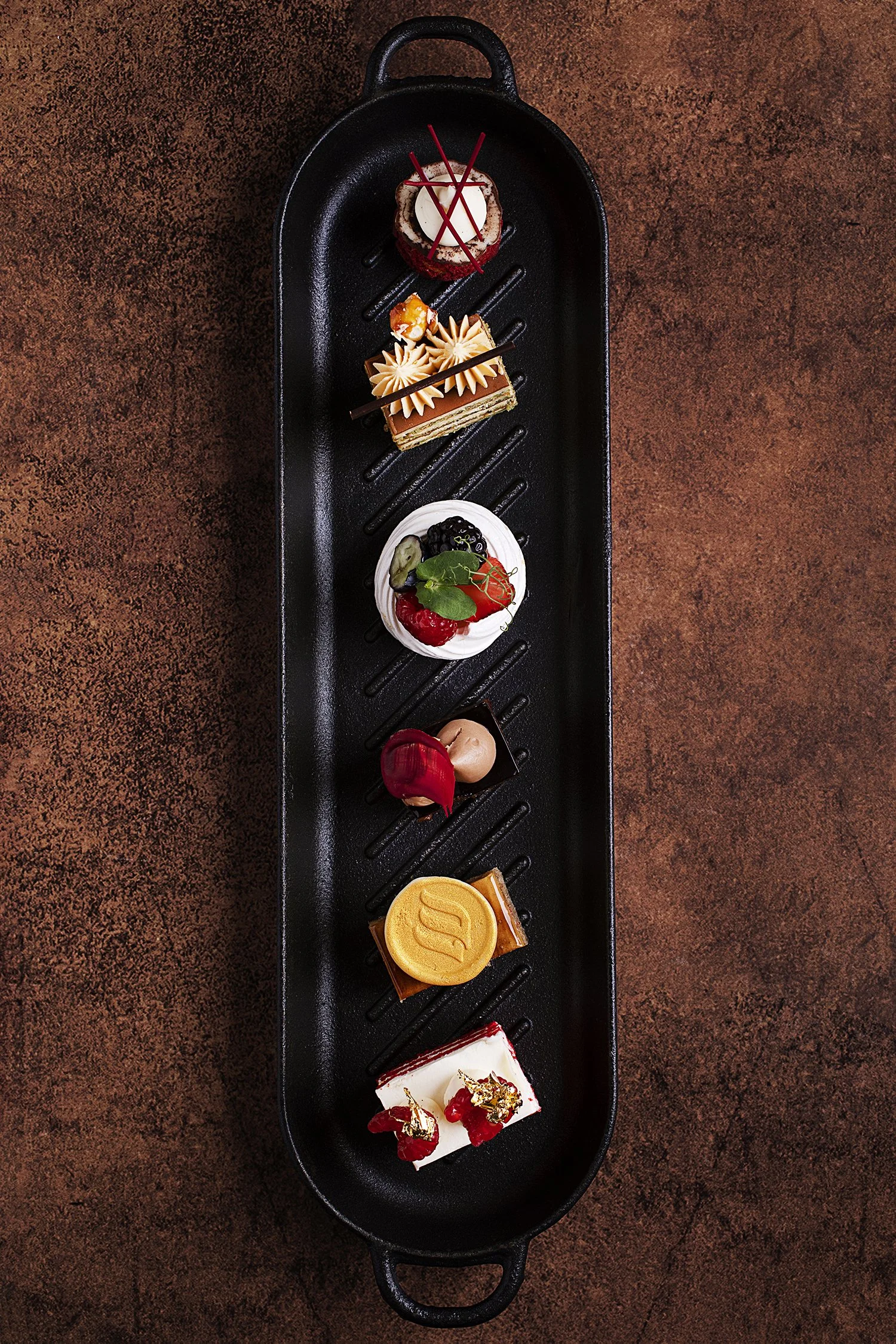 A black elongated serving platter holding six different small desserts, arranged vertically, on a brown textured surface. 