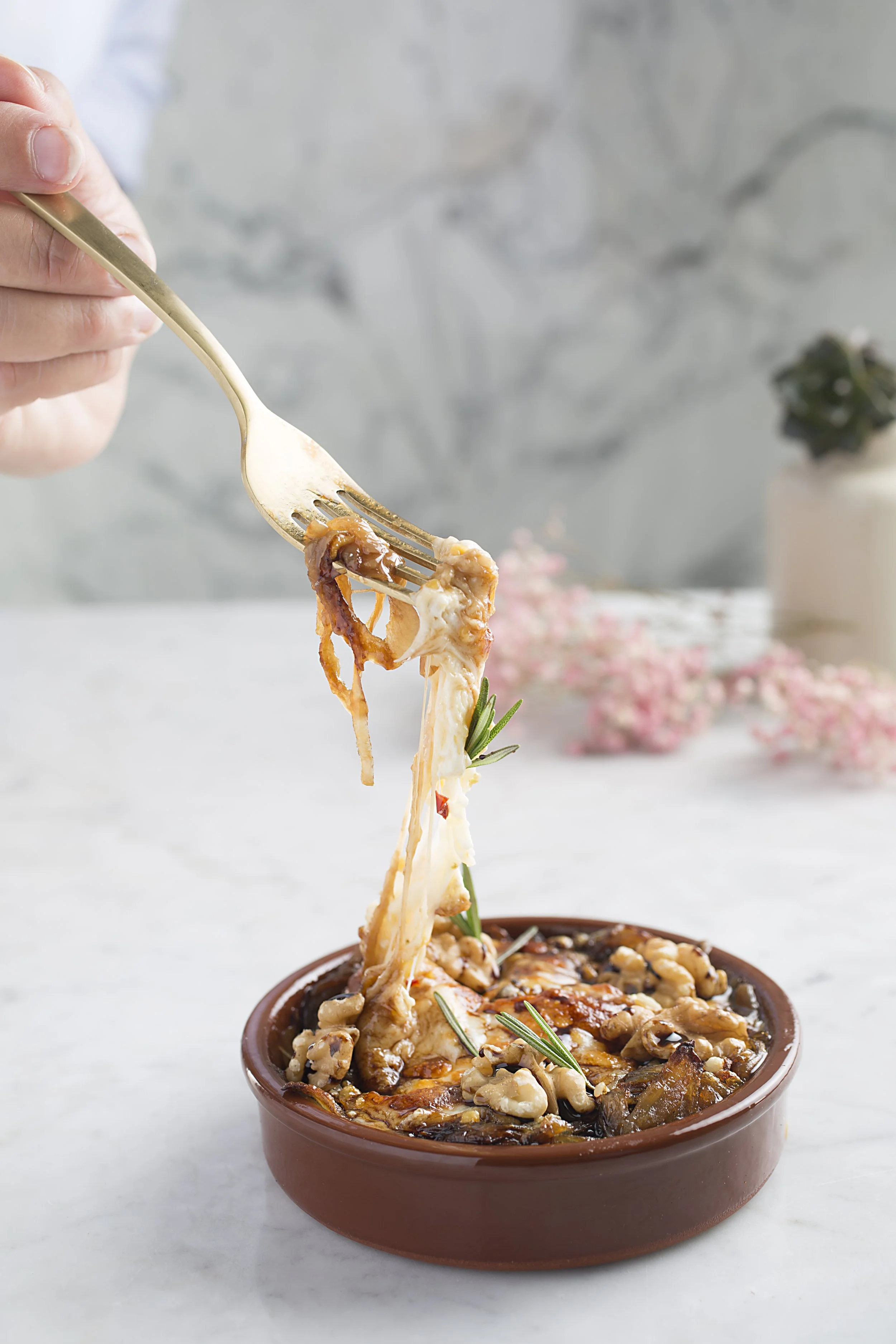 A piece of cheesy baked dish with nuts and herbs being lifted with a fork from a brown ceramic dish.