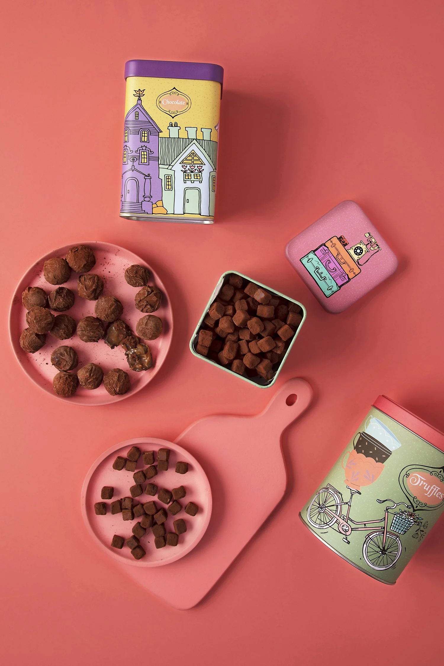 Assorted chocolates and truffles in pink and green tins, with some chocolates on pink plates, placed on a pink cutting board, all on a pink background. 