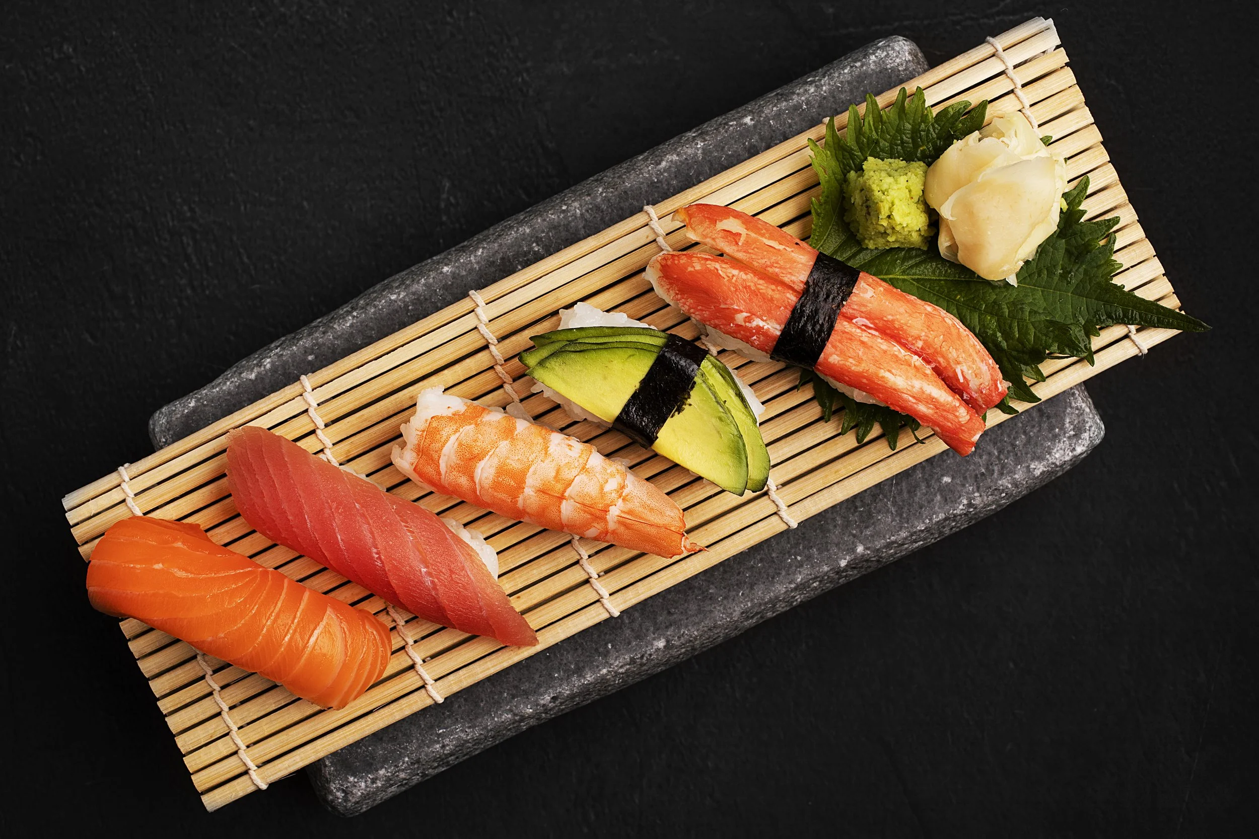 Assorted sushi pieces on a bamboo mat with wasabi, pickled ginger, and green leaf garnish on a black surface. Food photography in Kuwait
