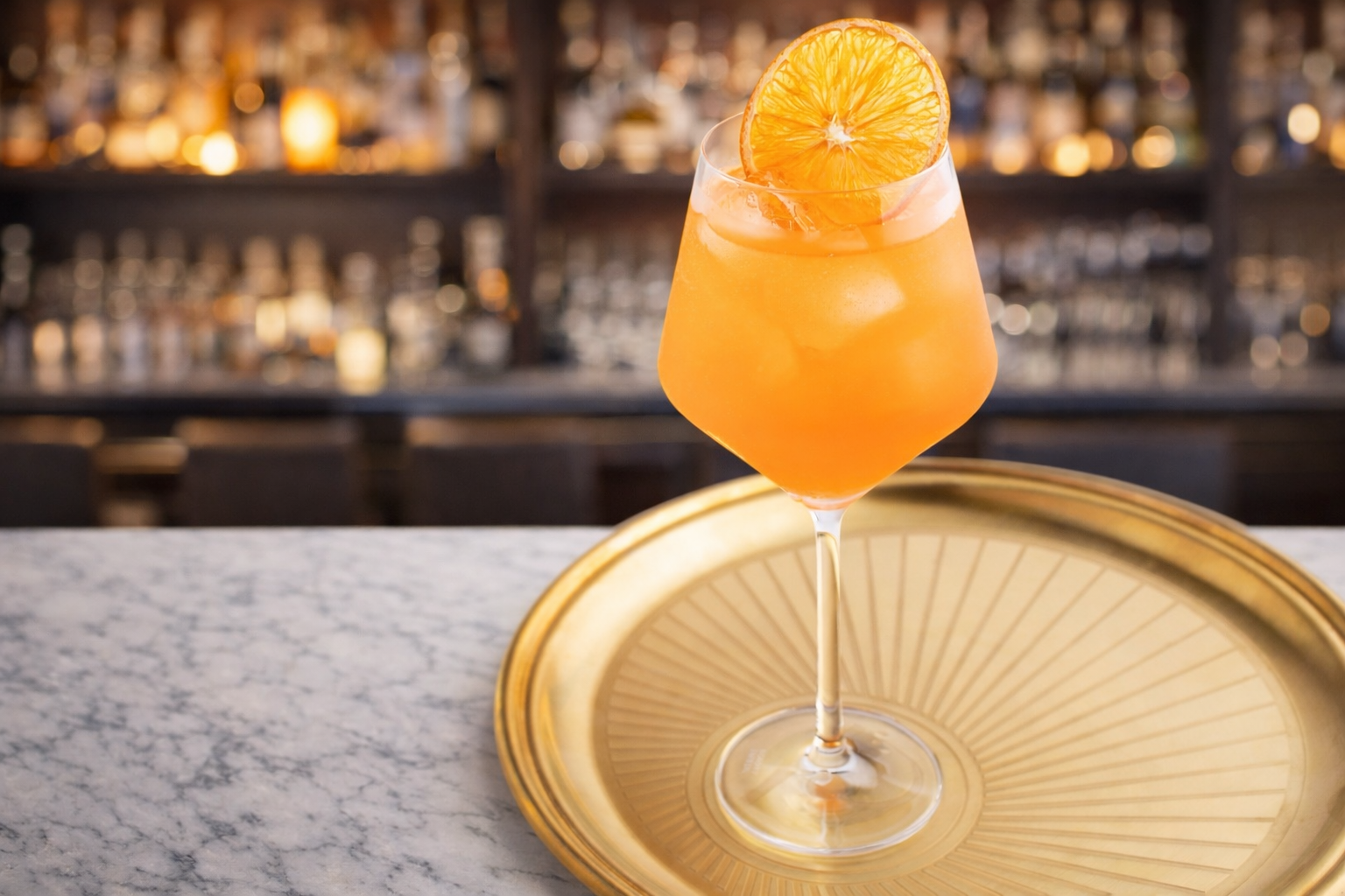 A cocktail drink with an orange slice garnish in a wine glass, placed on a gold tray on a marble countertop with a blurred bar background