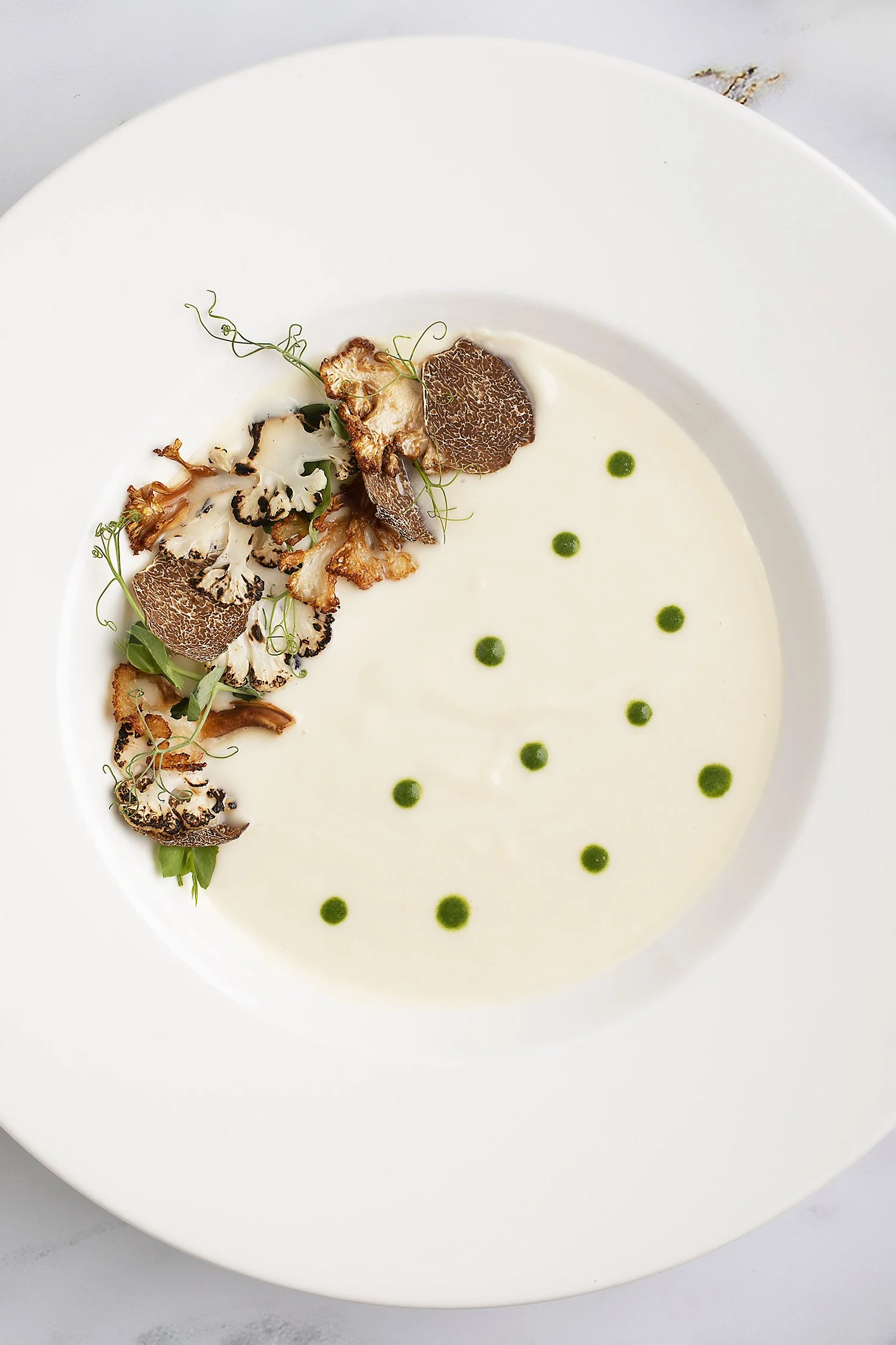 A white bowl of creamy cauliflower soup garnished with roasted mushrooms, microgreens, and green oil dots. 