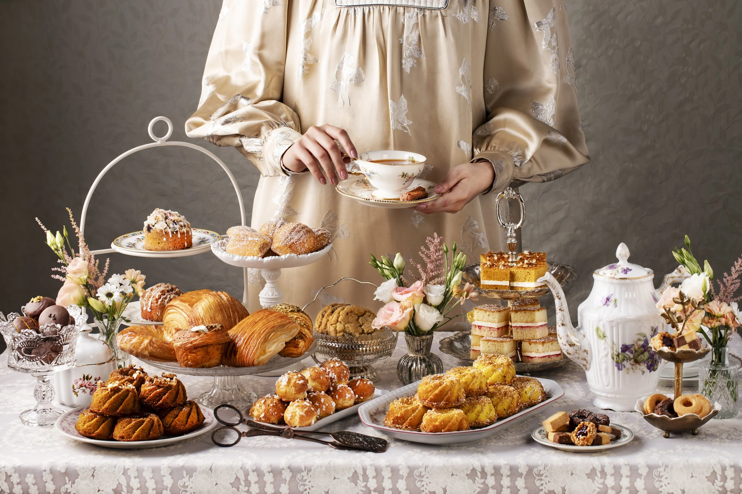 A woman in a beige floral dress serving tea at an ornate table filled with various pastries, cakes, and flowers for an afternoon tea. 