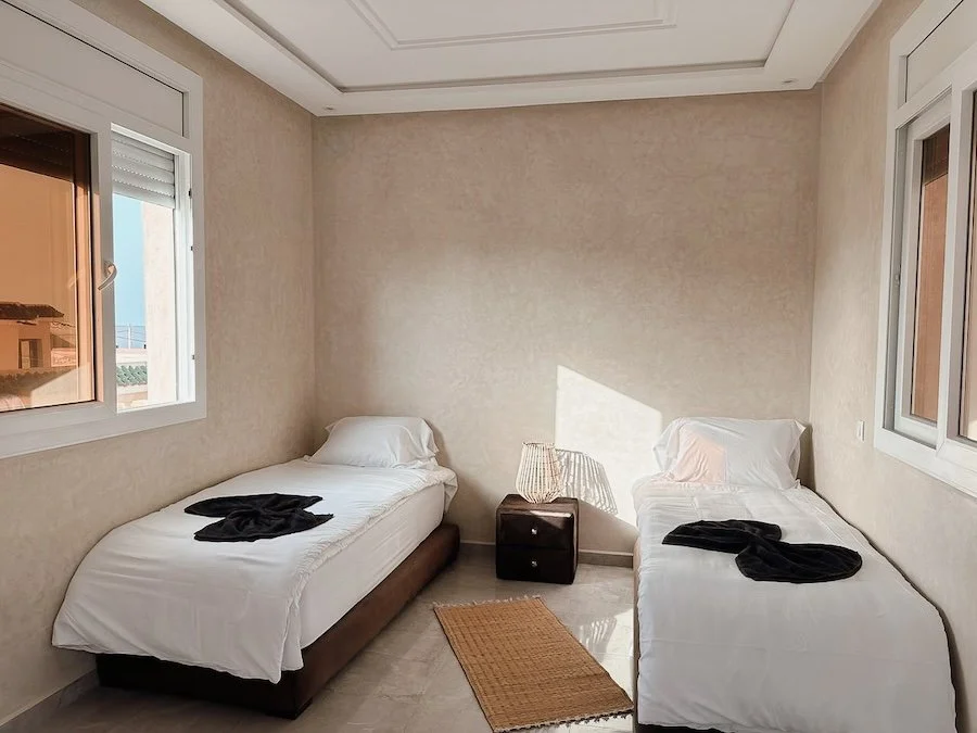 A bedroom with two single beds, each with white bedding and a black towel on top, a small nightstand with a lamp between the beds, and windows on two walls letting in natural light.