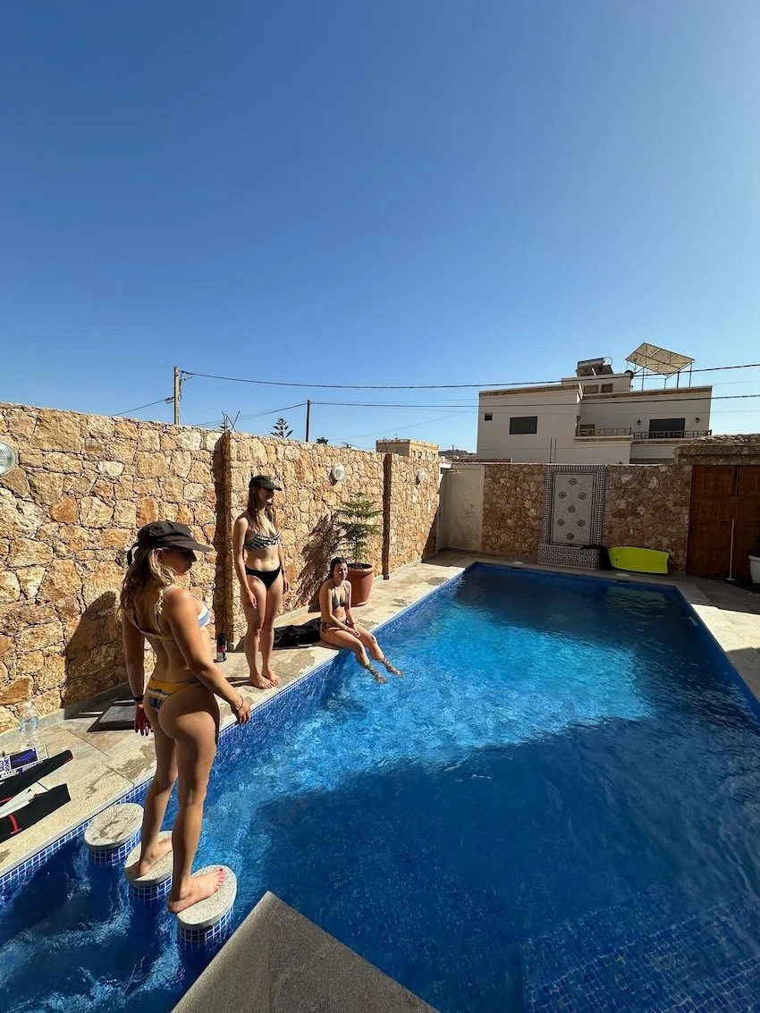 Three women in swimsuits by a private pool on a sunny day, one sitting with feet in the water, another standing, and the third stepping onto the pool edge.