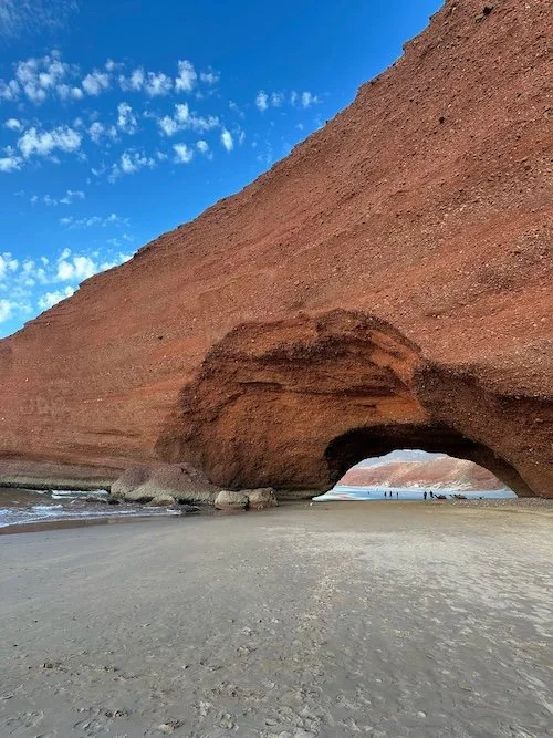 Red sandstone cliff with an arch on a sandy beach, blue sky with clouds in the background.