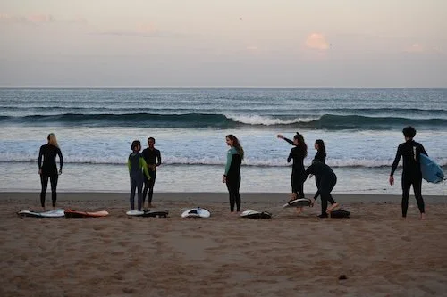Group of people preparing for a surf lesson on the beach at sunset with surfboards and wetsuits.