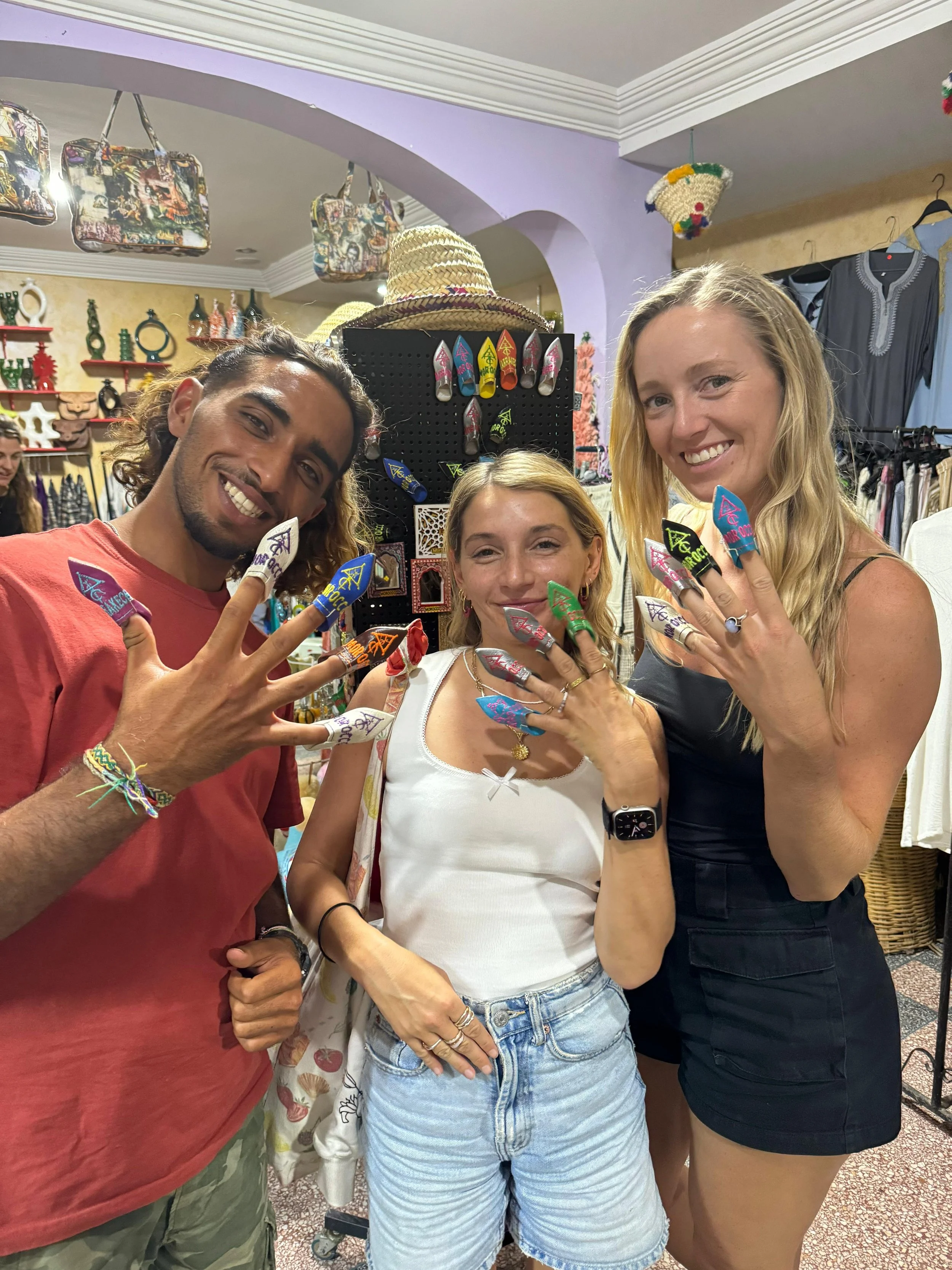 Three smiling women displaying colorful souvenir nails in a shop decorated with bags, hats, and souvenirs.