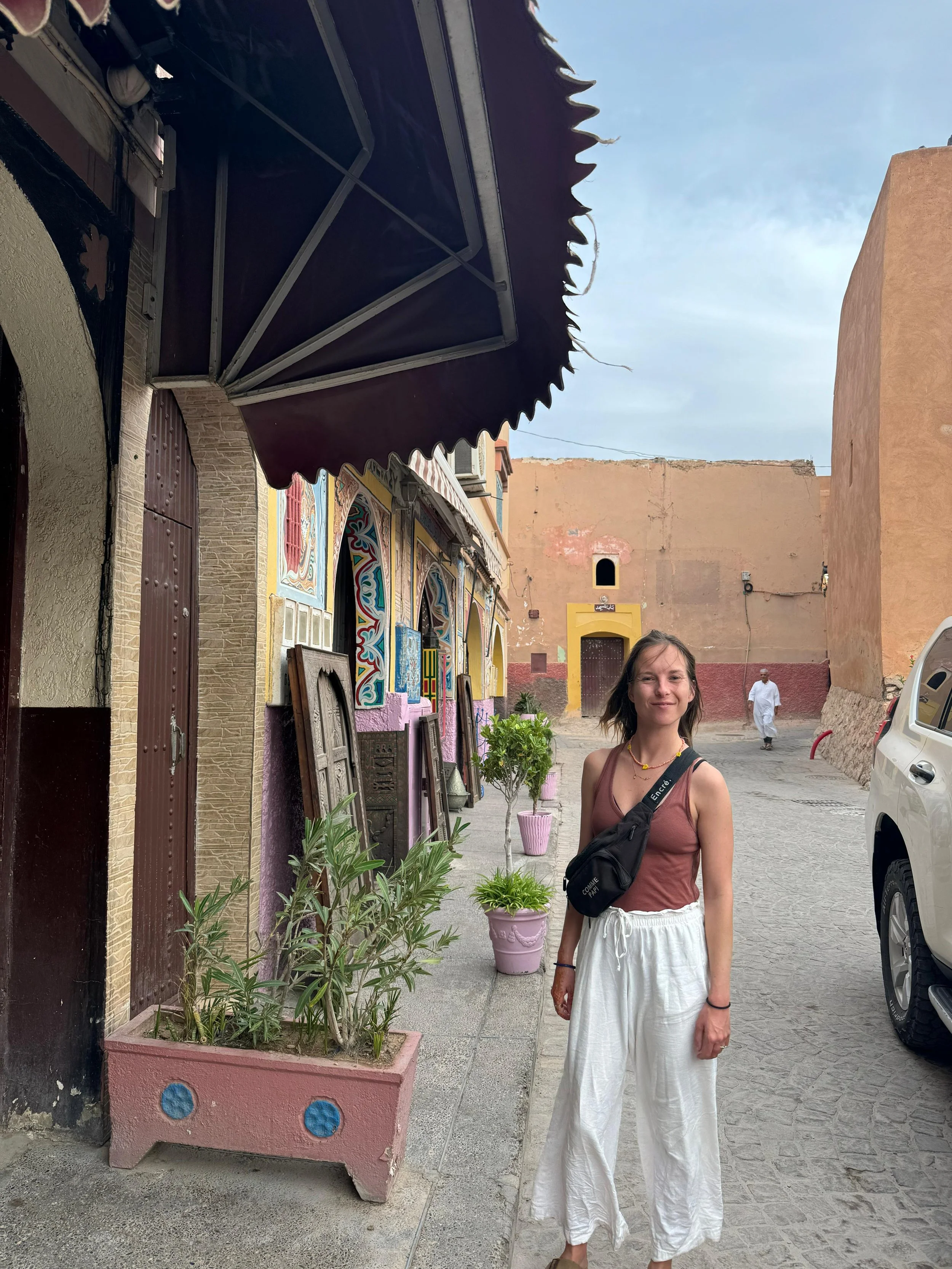 A woman standing on a sidewalk in front of colorful, decorated buildings with potted plants, a car, and a man in a white outfit in the background.