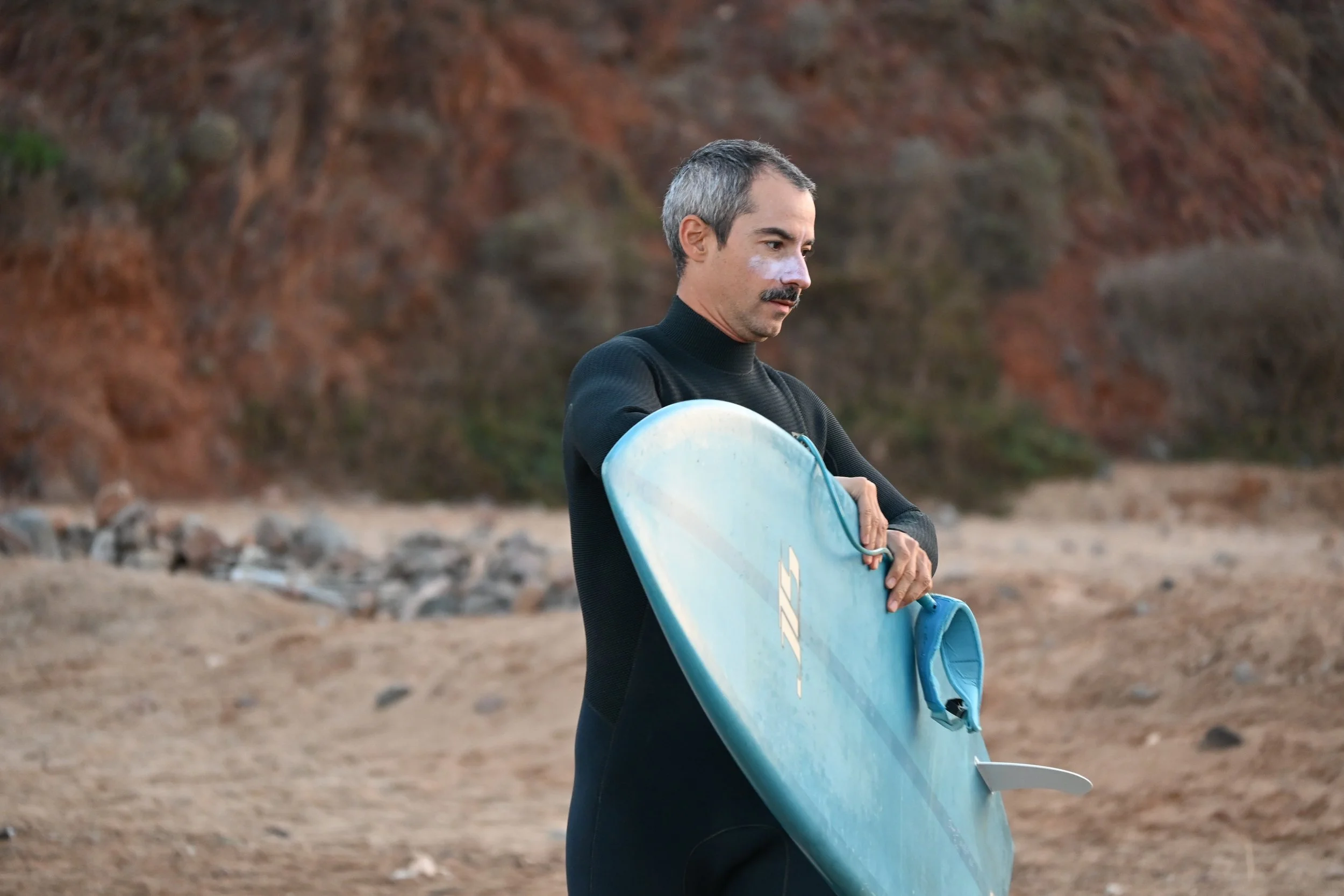 A man with gray and black hair, a mustache, and a black wetsuit holding a blue surfboard with a leash, standing on a sandy beach with rocky cliffs in the background.