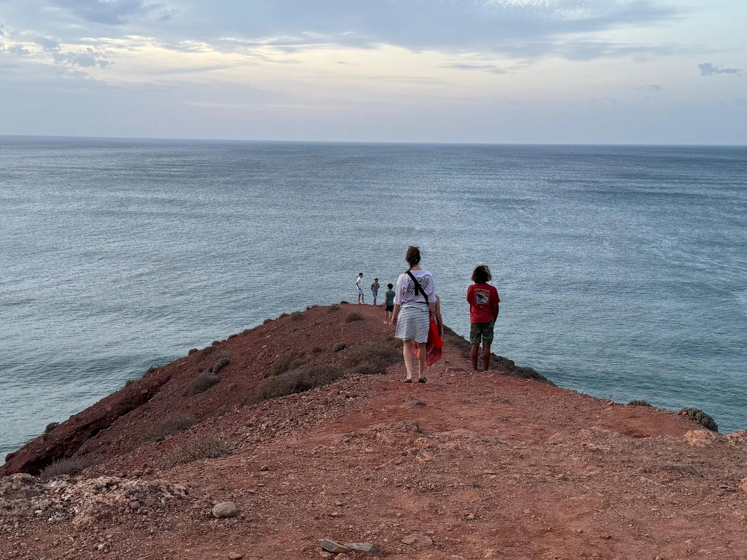 People walking on a dirt path along a rocky cliff overlooking the ocean at sunset.