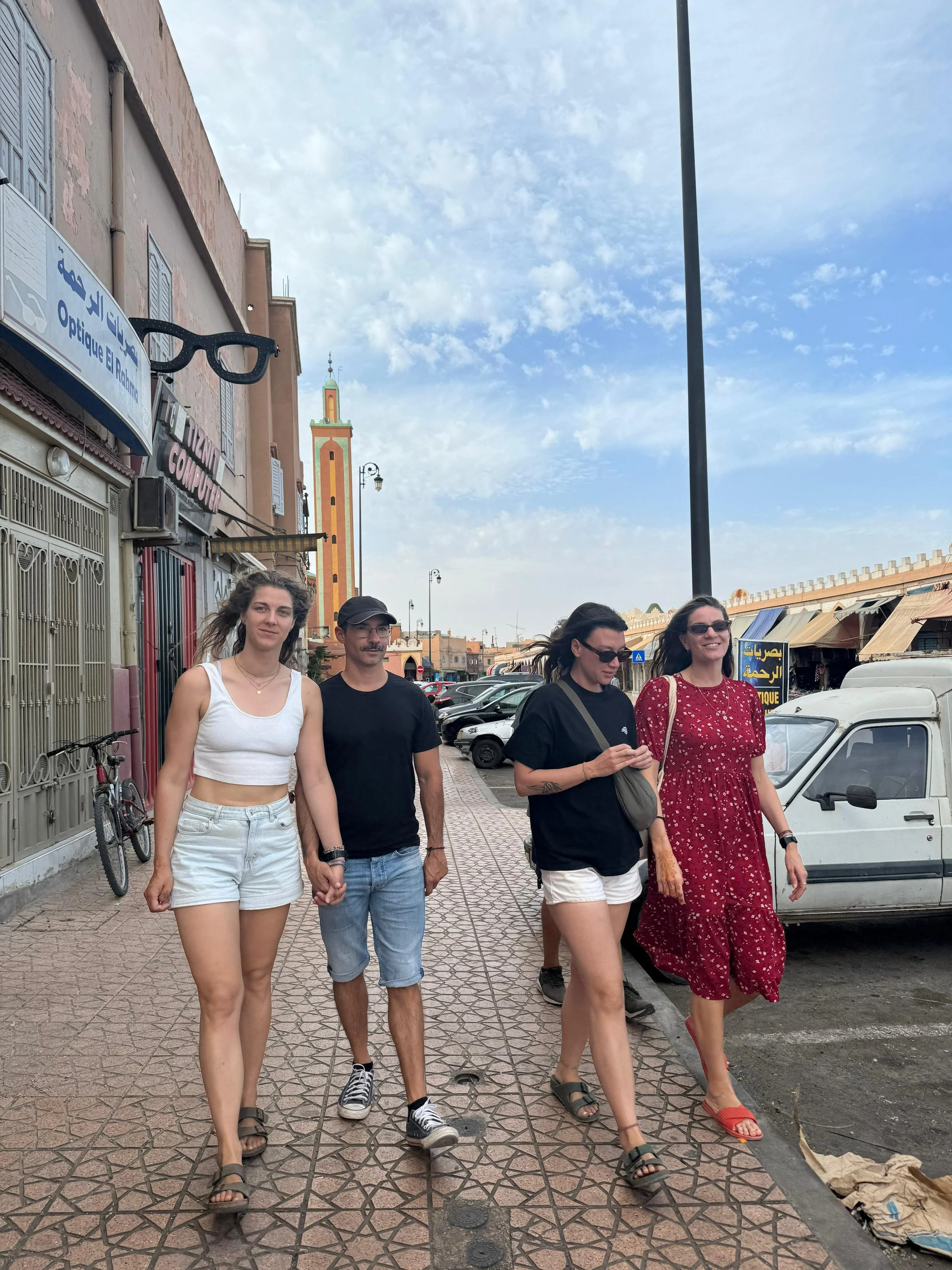 Four people walking on a sidewalk in a city, with a tall minaret in the background and parked cars on the side of the street.