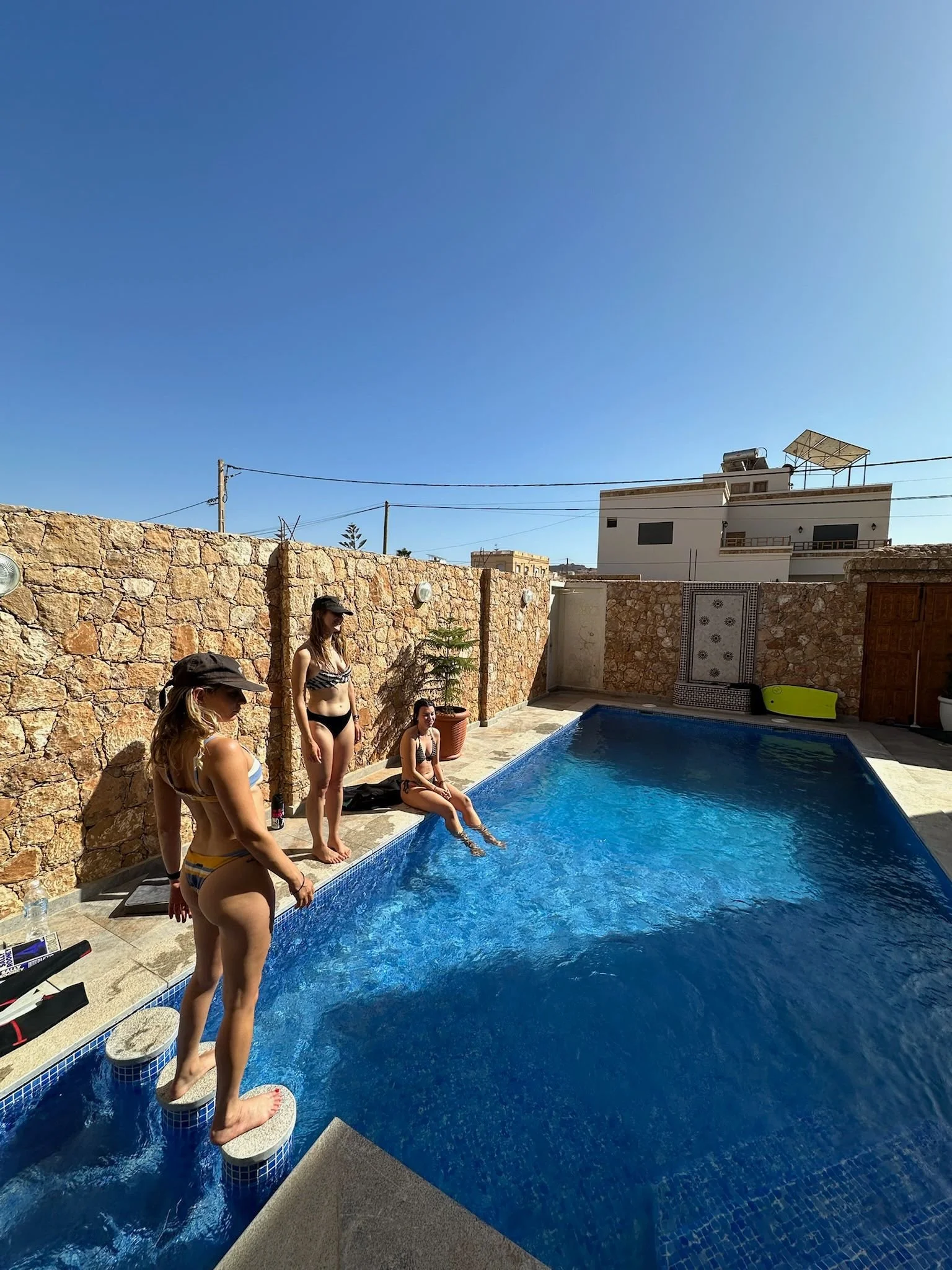 Three women in swimsuits near a small outdoor swimming pool with a stone wall and clear blue sky in the background.