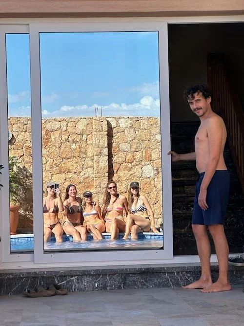 A man standing next to a sliding glass door, looking into the pool area where five women are sitting in a pool, smiling and posing. The outdoor scene includes a stone wall and a partly cloudy sky.