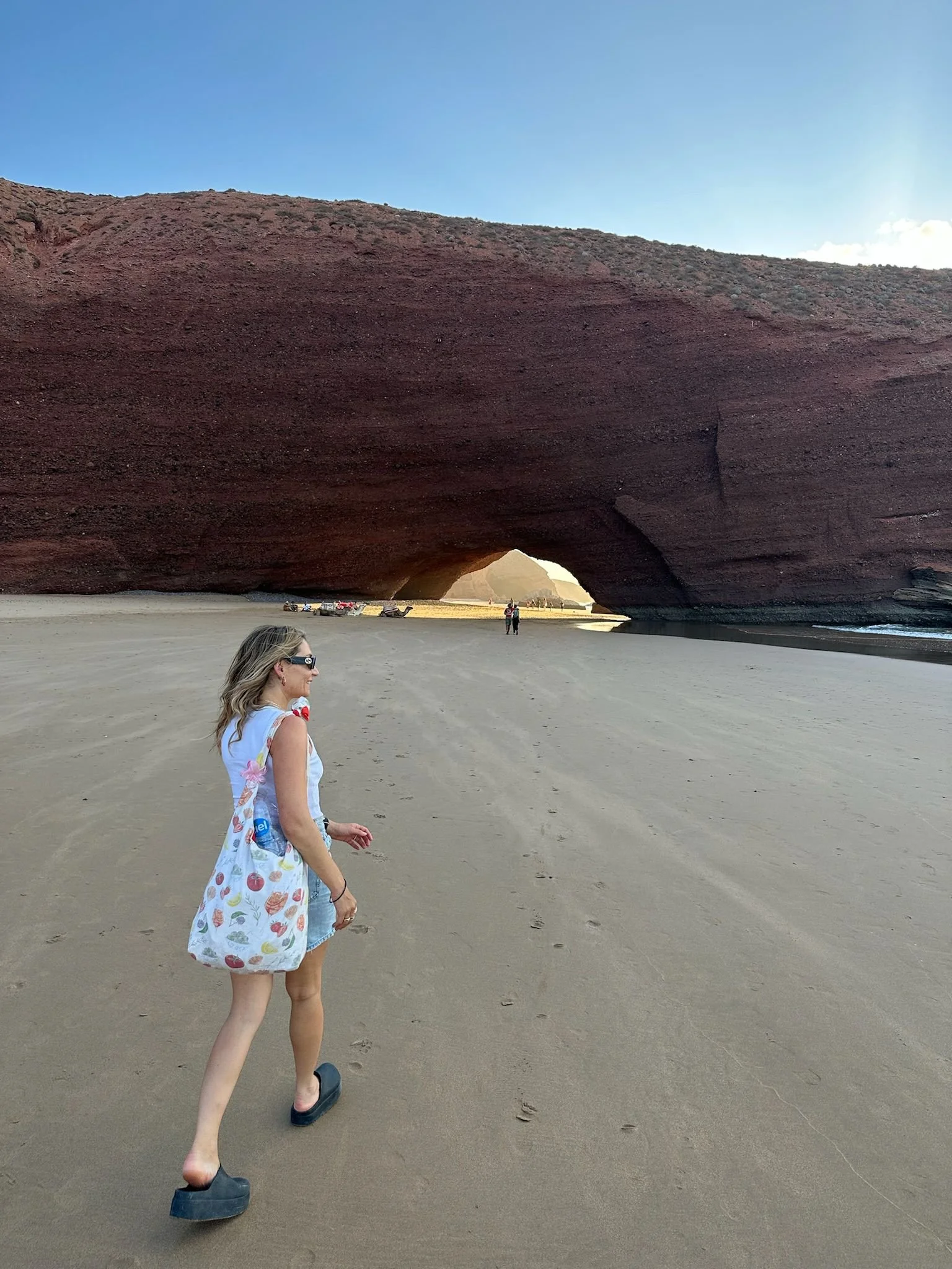 A woman walking on the sandy beach near a large, arched rock formation at a beach, with a few people in the distance and a clear blue sky overhead.