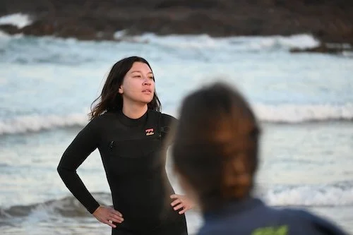 A woman in a black wetsuit standing on the beach near the ocean, with her hands on her hips, gazing into the distance, while another person is in the foreground with their back to the camera.