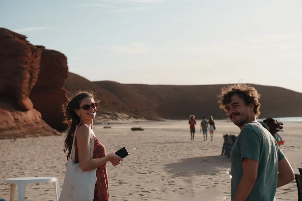 A woman and a man smile on a beach with rocky cliffs in the background. The woman is holding a phone and wearing sunglasses, while the man looks back toward the camera. Other people are walking in the distance.