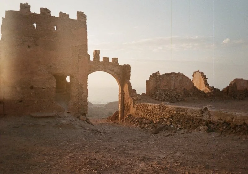 Ruins of an ancient stone fortress with an arched gateway, overgrown stone walls, and a dirt ground, under a cloudy sky at sunset.