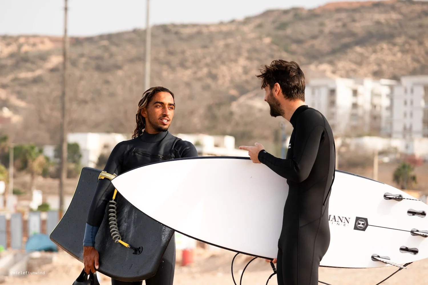 Two men in wetsuits having a conversation on the beach, one holding a surfboard and the other holding a surfboard and a wetsuit bag, with sand, buildings, and hills in the background.
