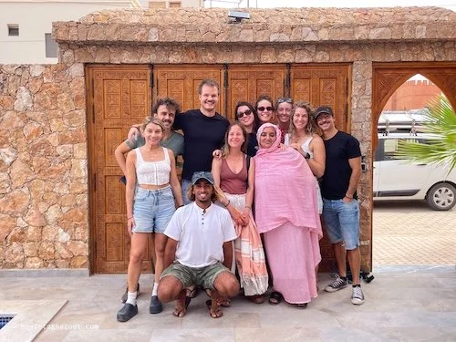 A group of nine people, including a woman wrapped in pink fabric, standing in front of a stone wall with a wooden door, in an outdoor setting with a parked car and palm tree in the background.