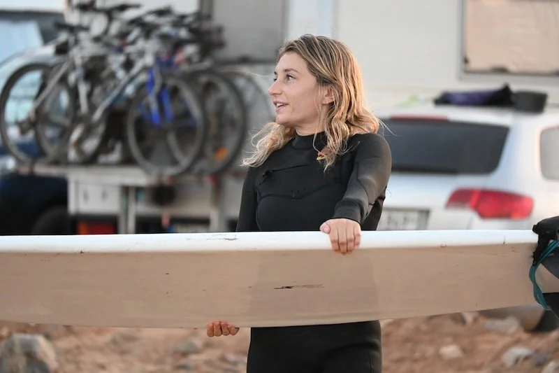 A blonde girl ready to get surfing wearing a wetsuit and carrying a white surfboard to the beach
