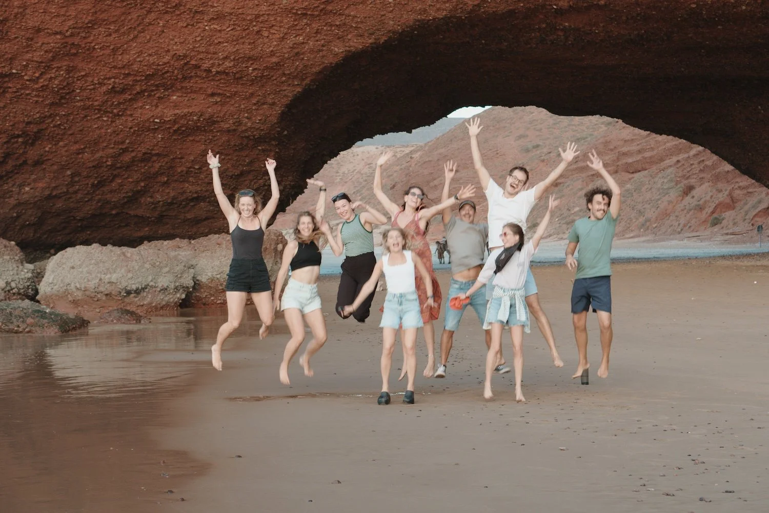 Group of ten friends jumping in the air on a sandy beach under a large red rock arch formation.