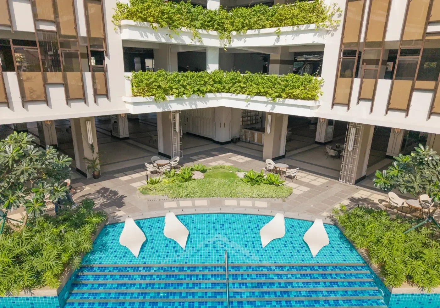 View of a rooftop pool with three white modern lounge chairs, surrounded by greenery, with a multi-story building with balconies and large glass windows in the background.