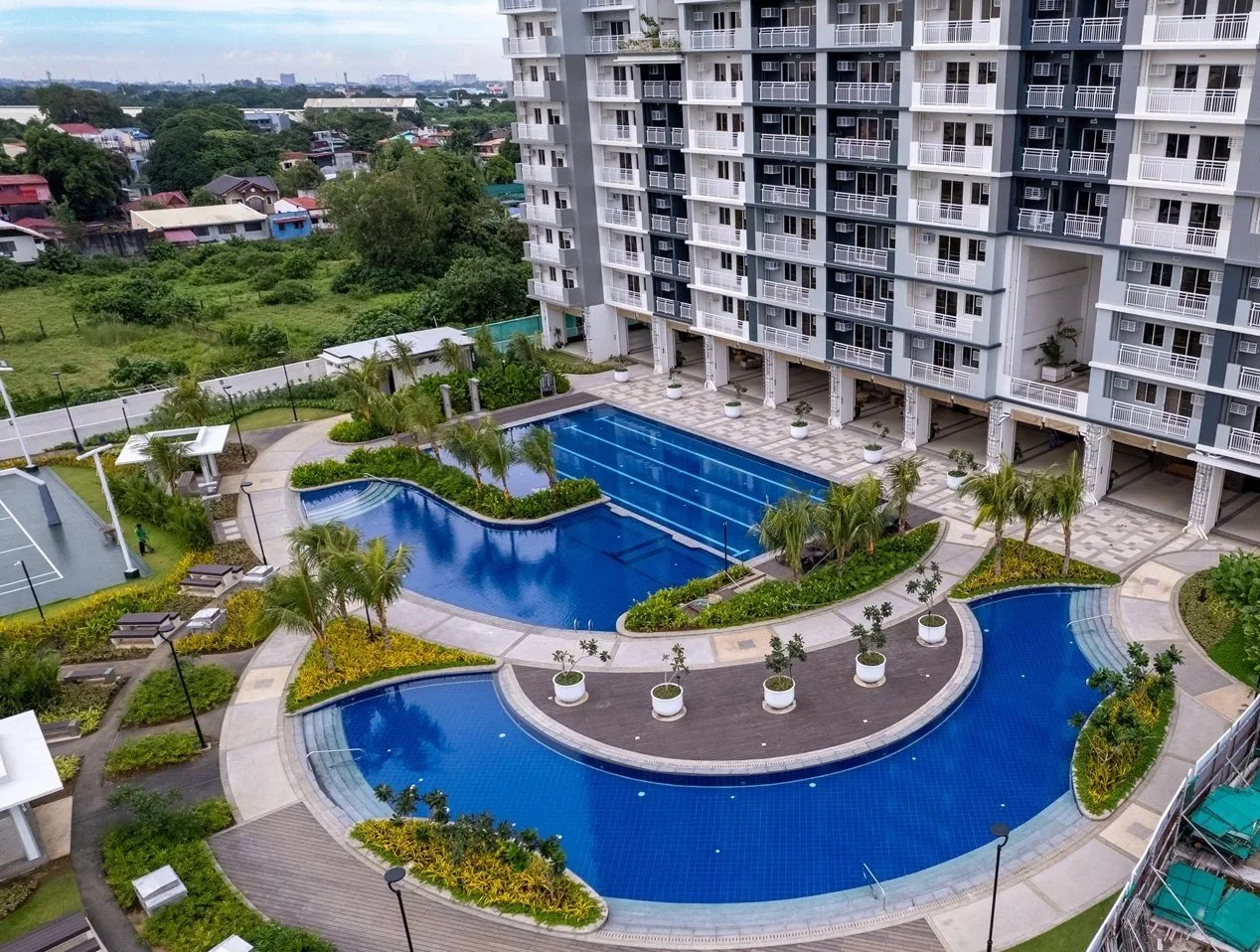 An aerial view of a swimming pool and surrounding landscape in a high-rise apartment complex, with greenery and residential houses in the background.
