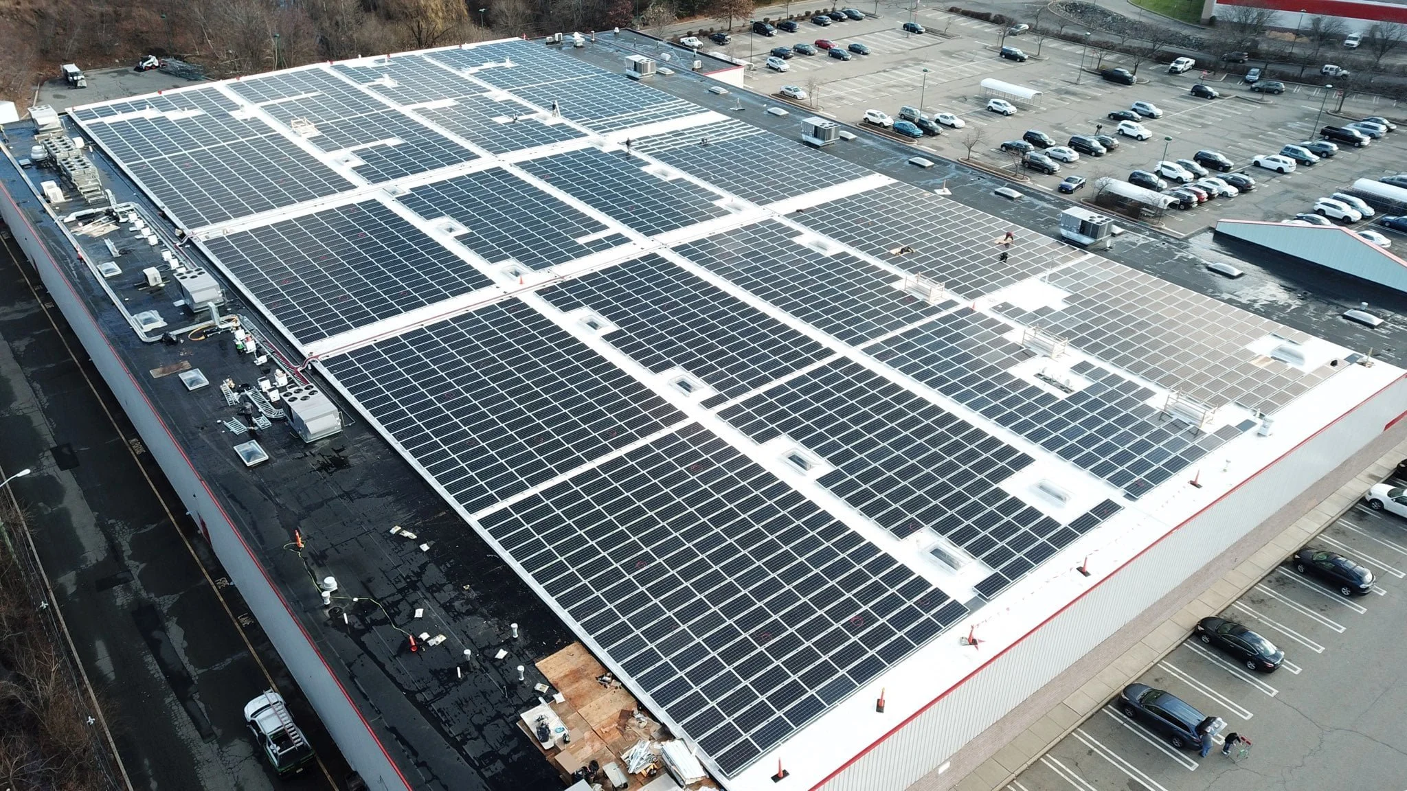 Aerial view of a large commercial building with a flat roof covered in numerous solar panels. Surrounding the building are parking lots filled with cars.