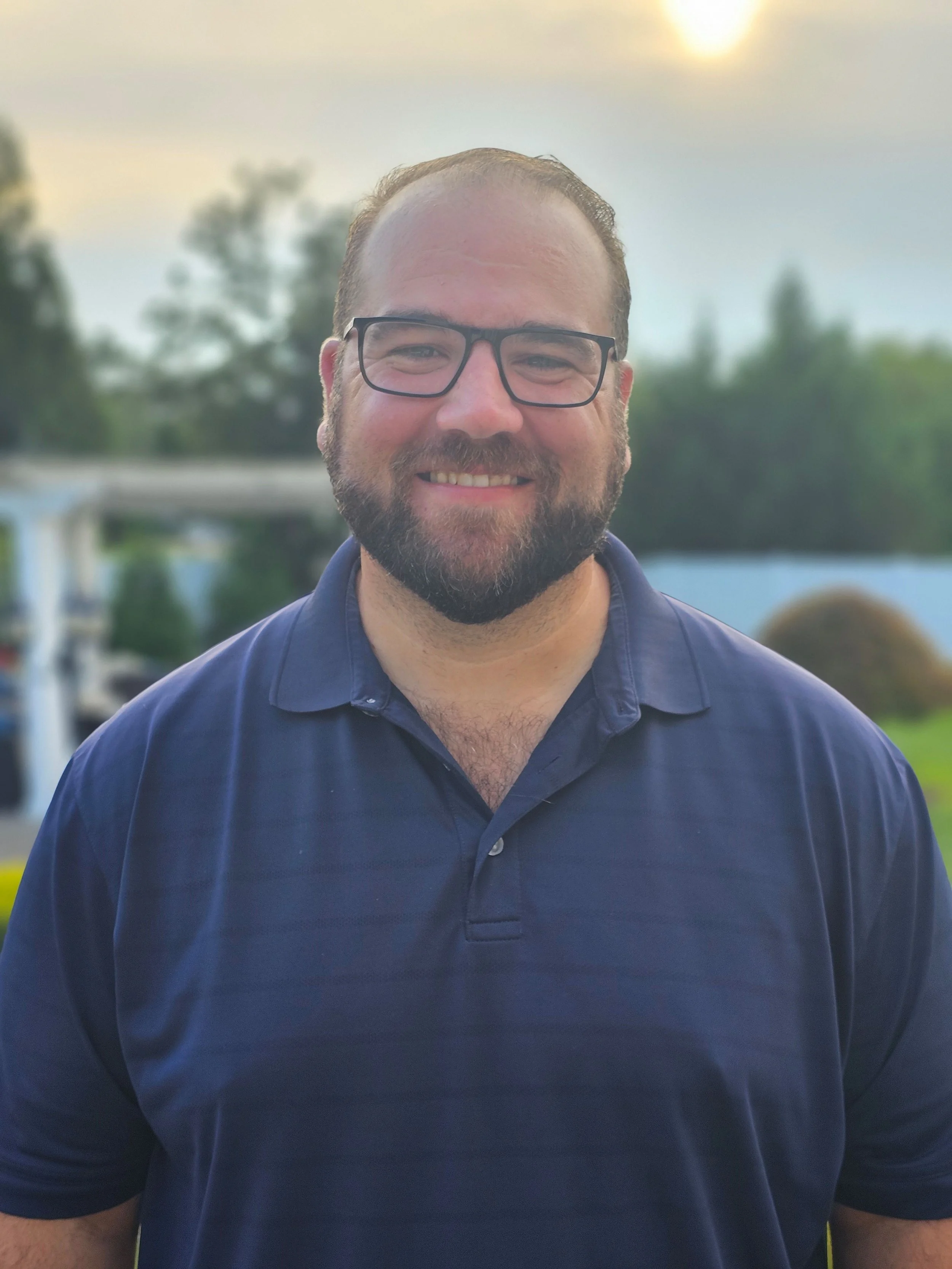A smiling man with glasses, a beard, and short hair stands outdoors during sunset, wearing a navy blue polo shirt, with trees and a building in the background.