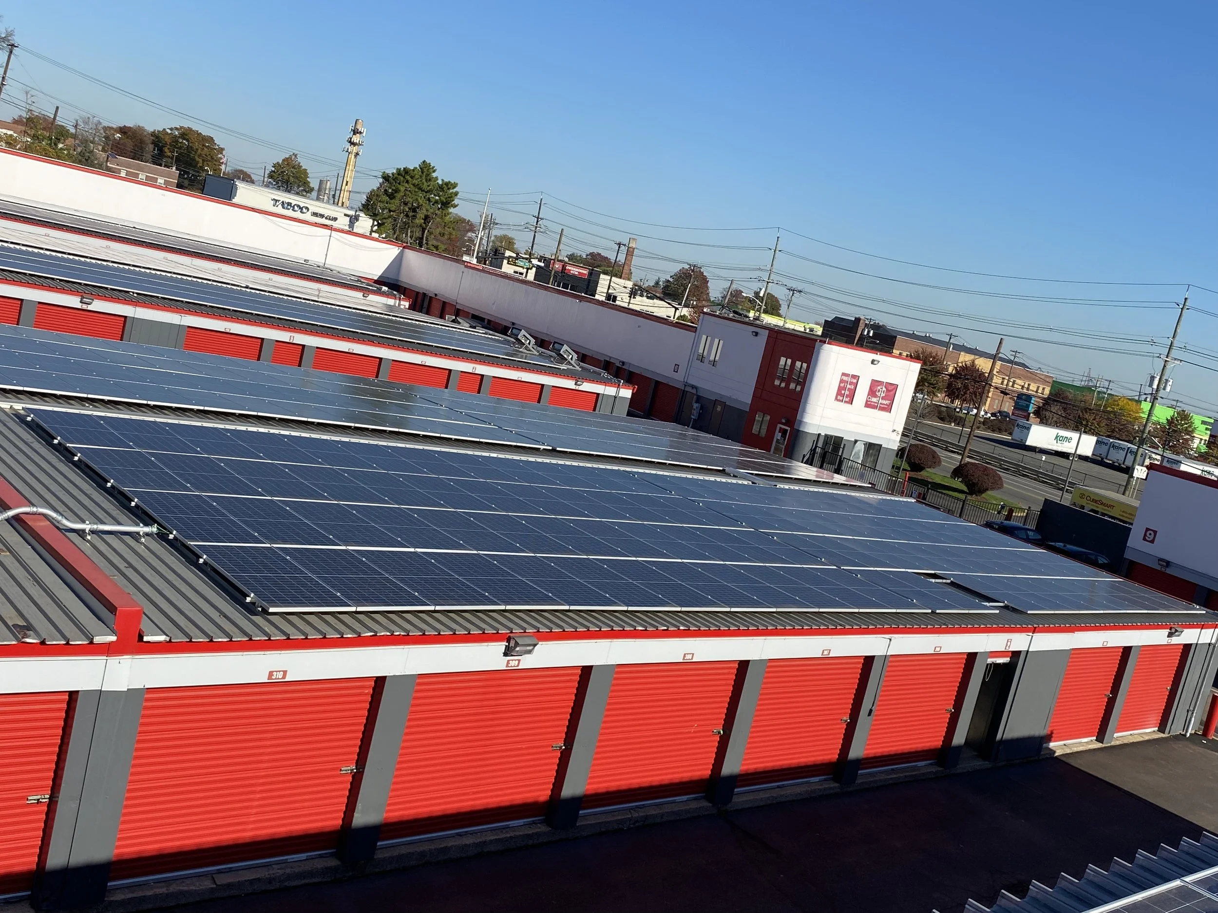 Multiple rows of solar panels installed on the roof of a storage facility with red roll-up doors, surrounded by trees, power lines, and commercial buildings under a clear blue sky.