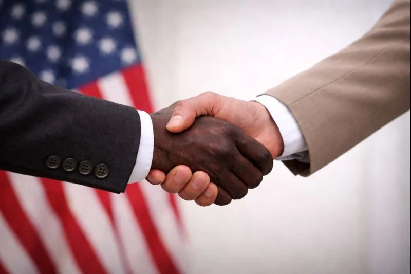 Two people shaking hands, one in a dark suit and the other in a light suit, with an American flag in the background.