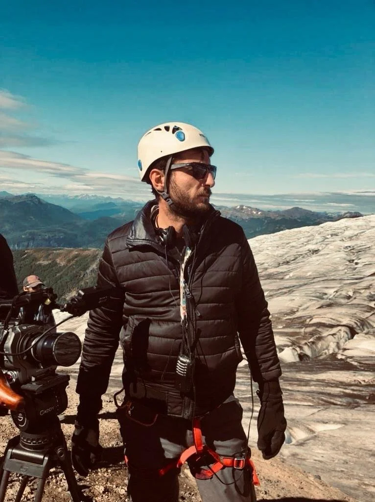 Man wearing a white helmet, sunglasses, black jacket, and gloves standing on a glacier with snowy mountains and a blue sky in the background.