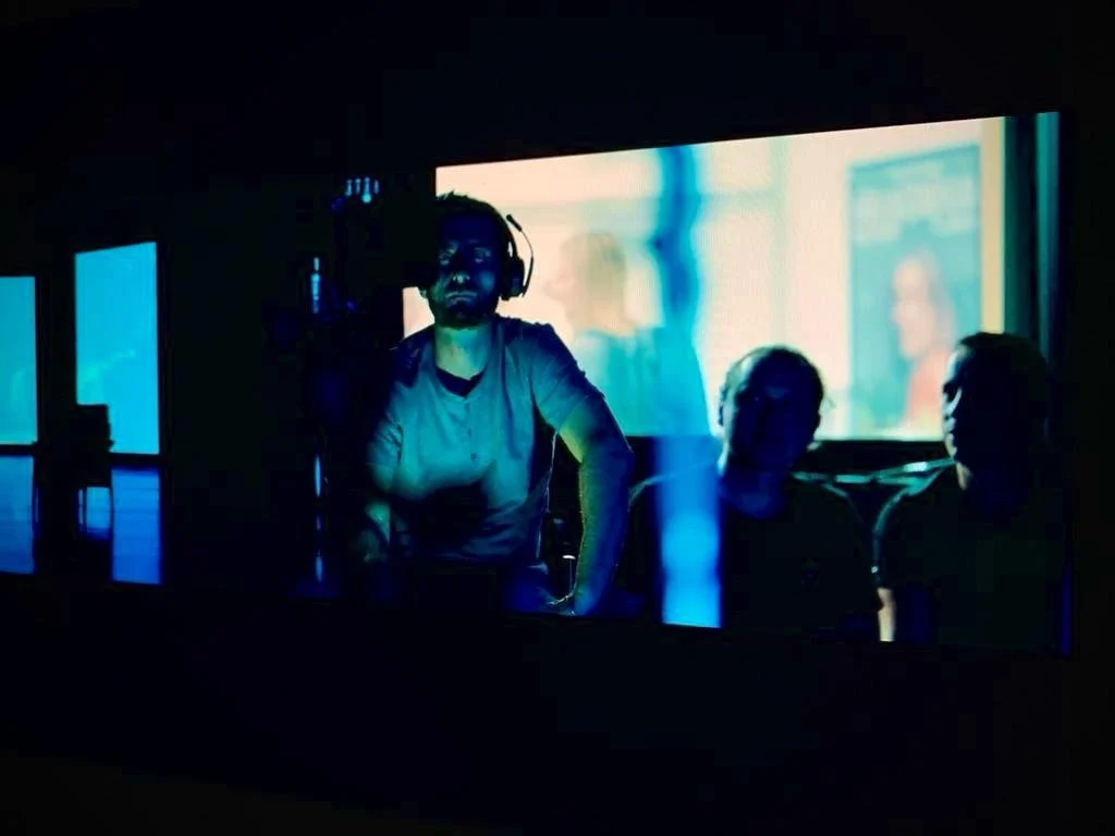 A person wearing headphones and a t-shirt, possibly a DJ, in a dark room with large screen displays in the background showing blurred images, with two other people sitting nearby.