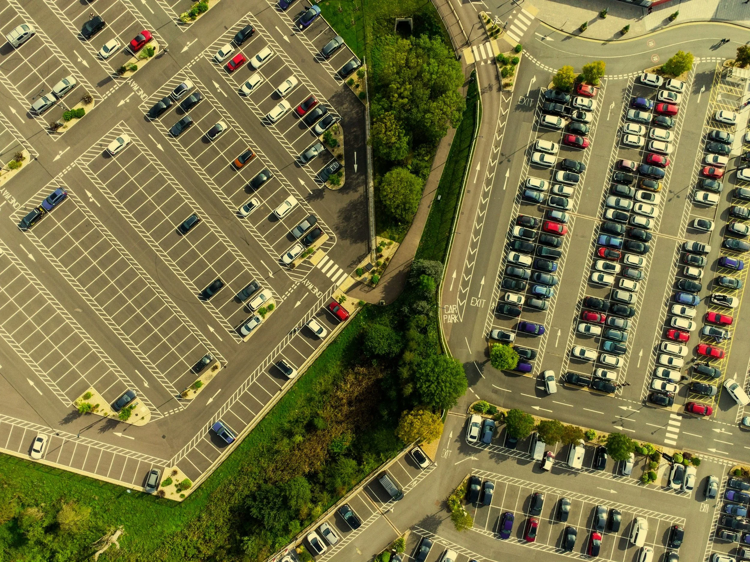 An aerial view of a large parking lot filled with cars, with clearly marked parking spaces, drive lanes, and greenery along the edges.