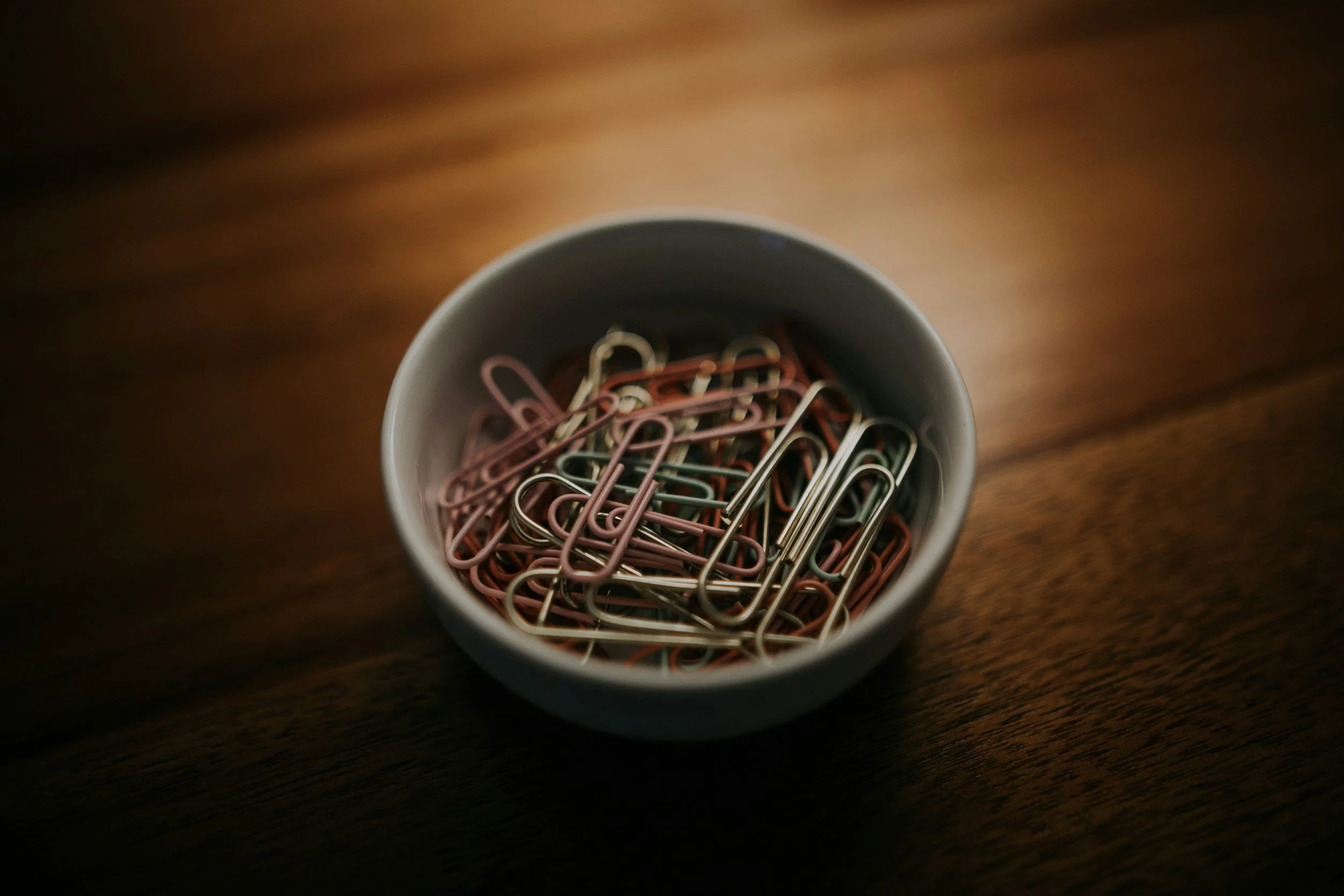 A white bowl filled with assorted pink and silver paperclips on a wooden surface.