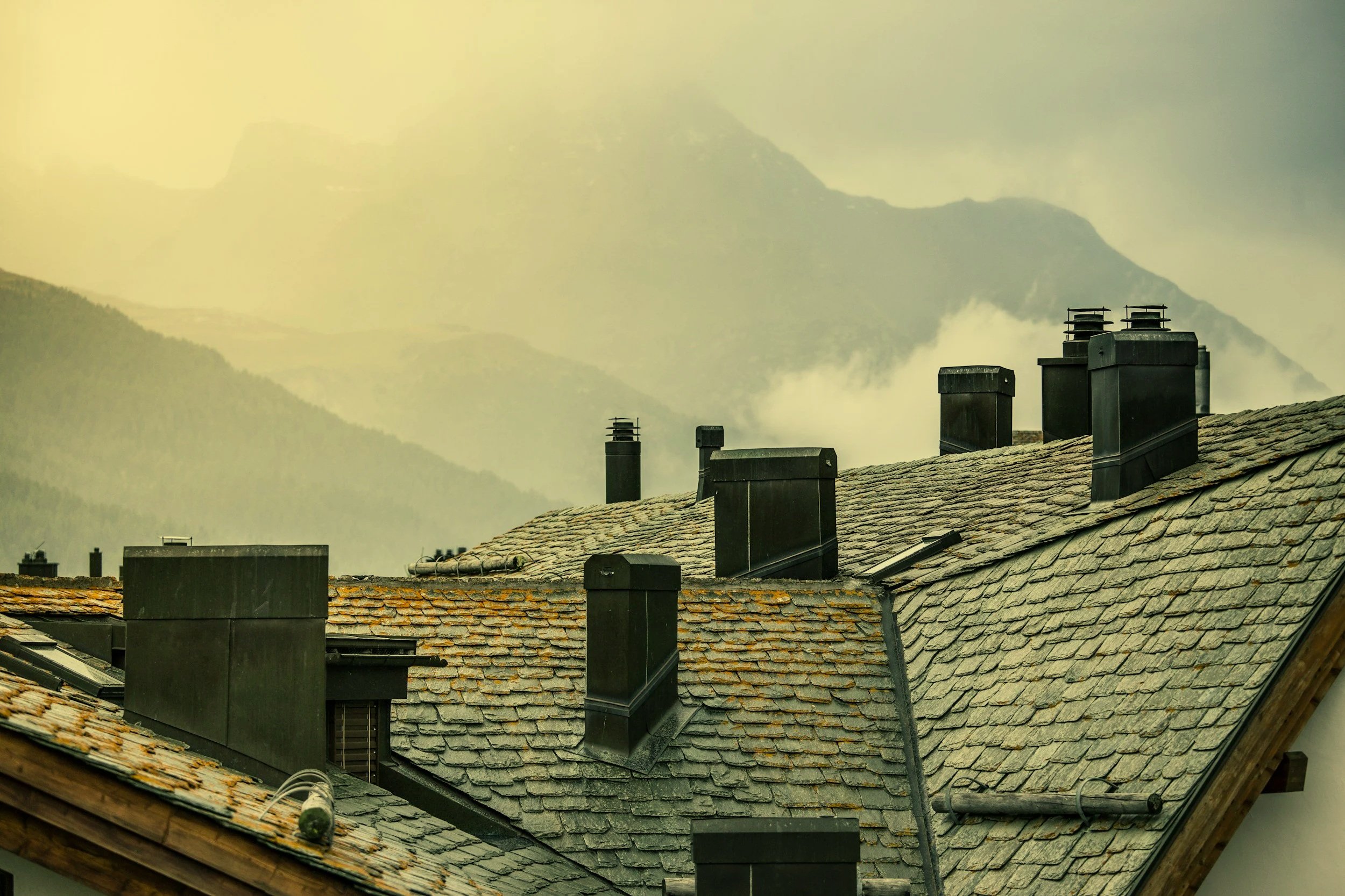 Several black chimneys on a gray tiled roof with mountains and cloudy sky in the background.