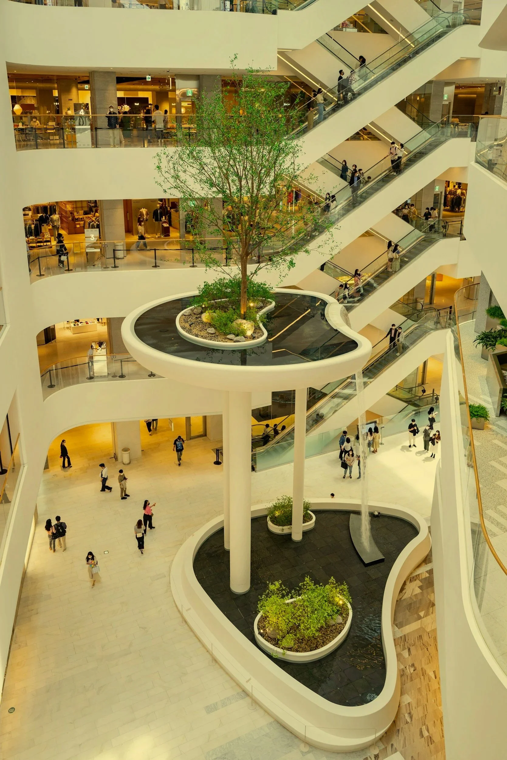 Interior of a modern shopping mall featuring multiple floors with escalators, a large central tree, decorative water features, and shoppers walking around.