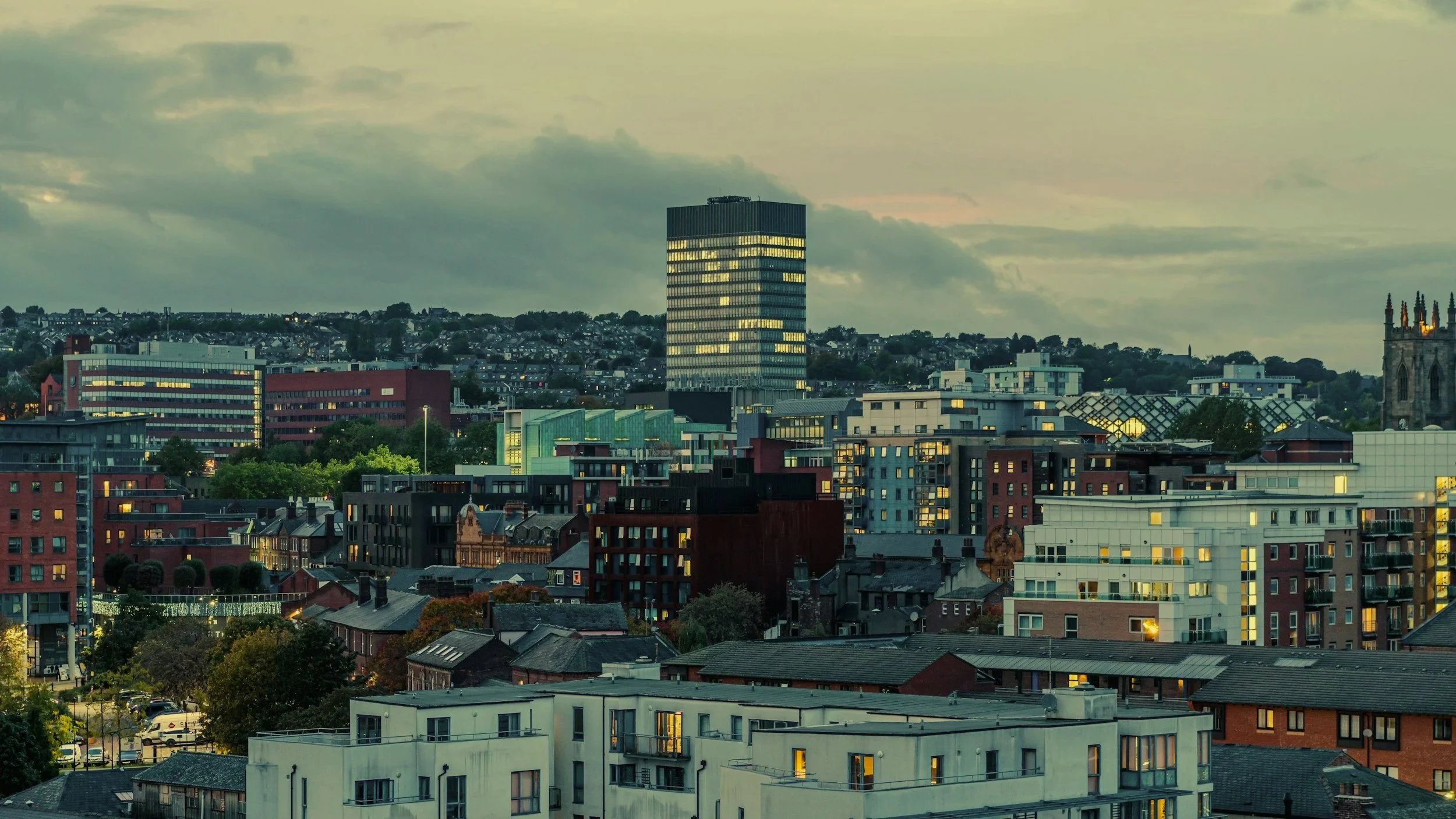 Cityscape during twilight with a mix of modern and historic buildings, some with illuminated windows, and a cloudy sky overhead.