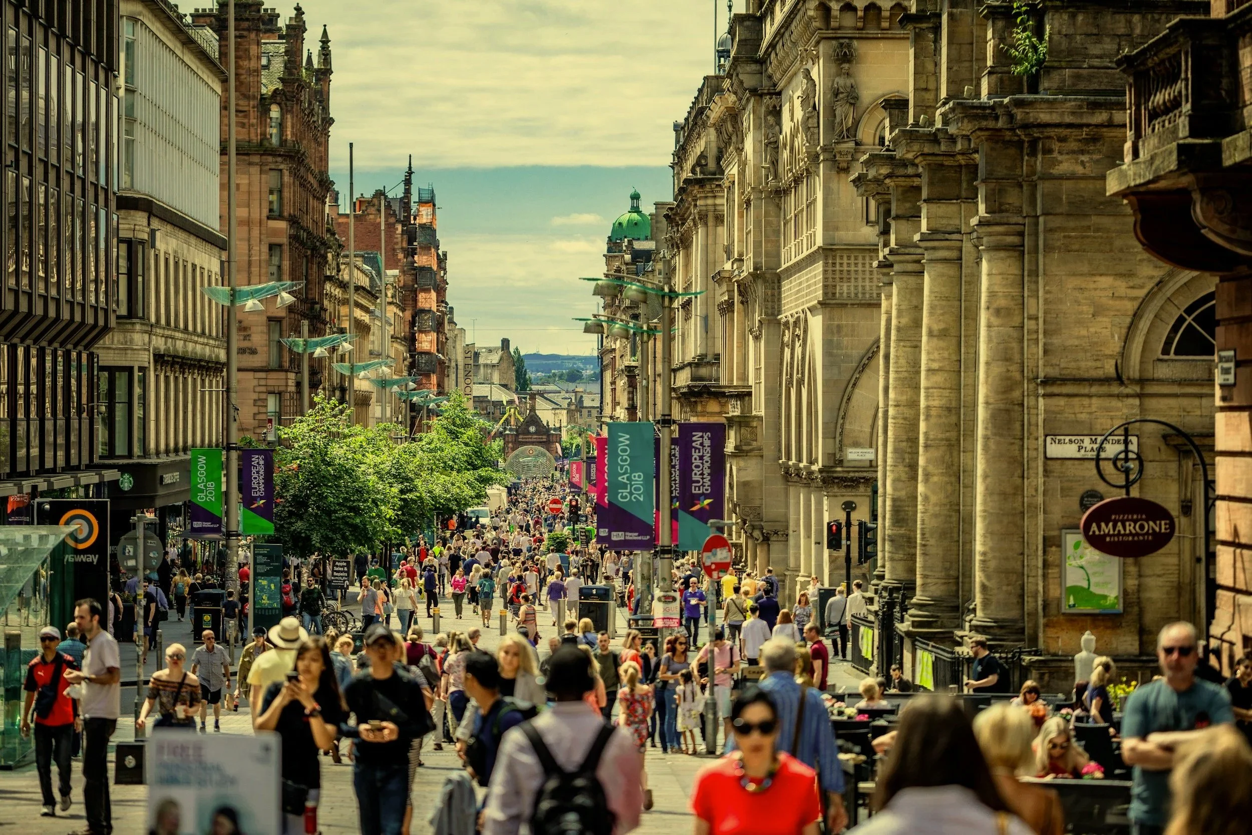 Busy city street filled with pedestrians, surrounded by historic buildings and banners advertising the Glasgow 2018 European Championships.