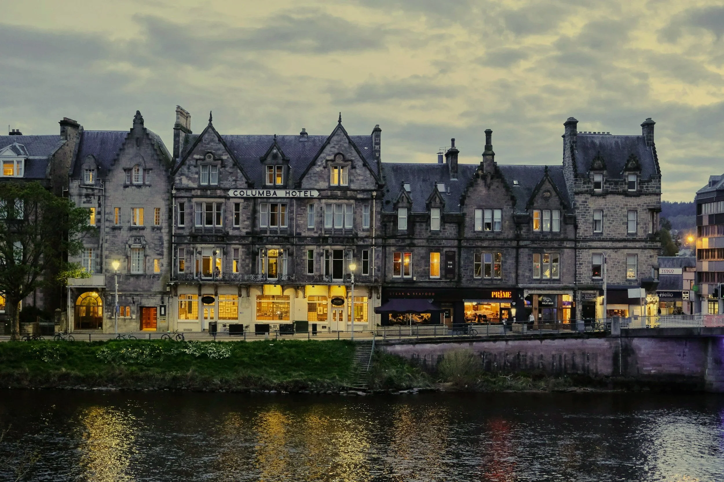 Historic stone building along a river with warm lit windows and a cloudy evening sky.