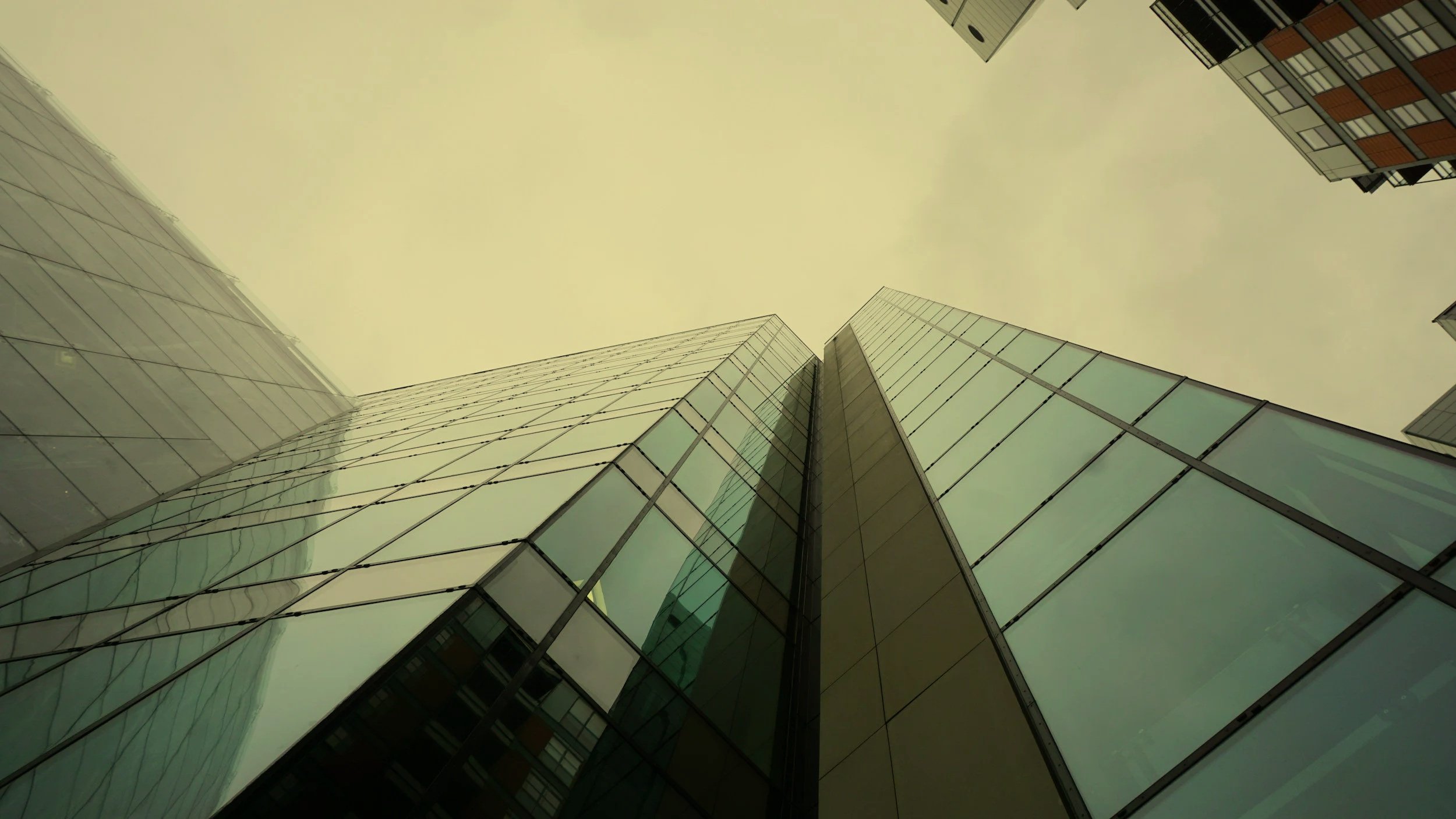 Looking up at modern glass skyscrapers from the ground, with a cloudy, yellowish sky above.
