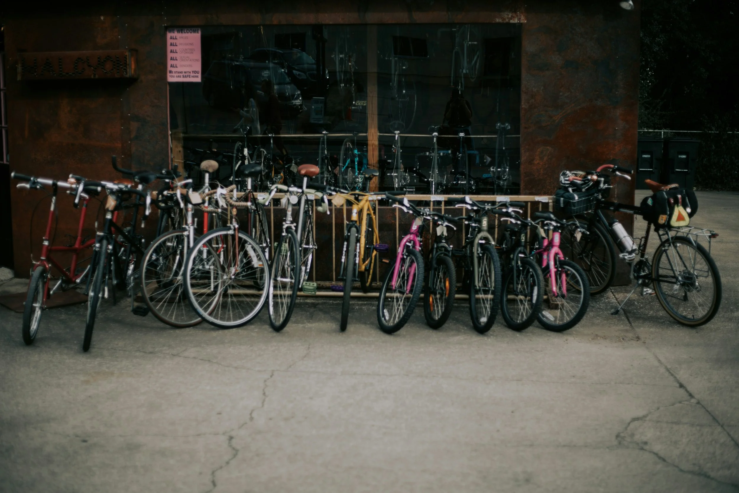 Bicycle rack outside a bike shop with various bikes parked in front, including pink, yellow, black, and red bikes, some with baskets and baby seats.